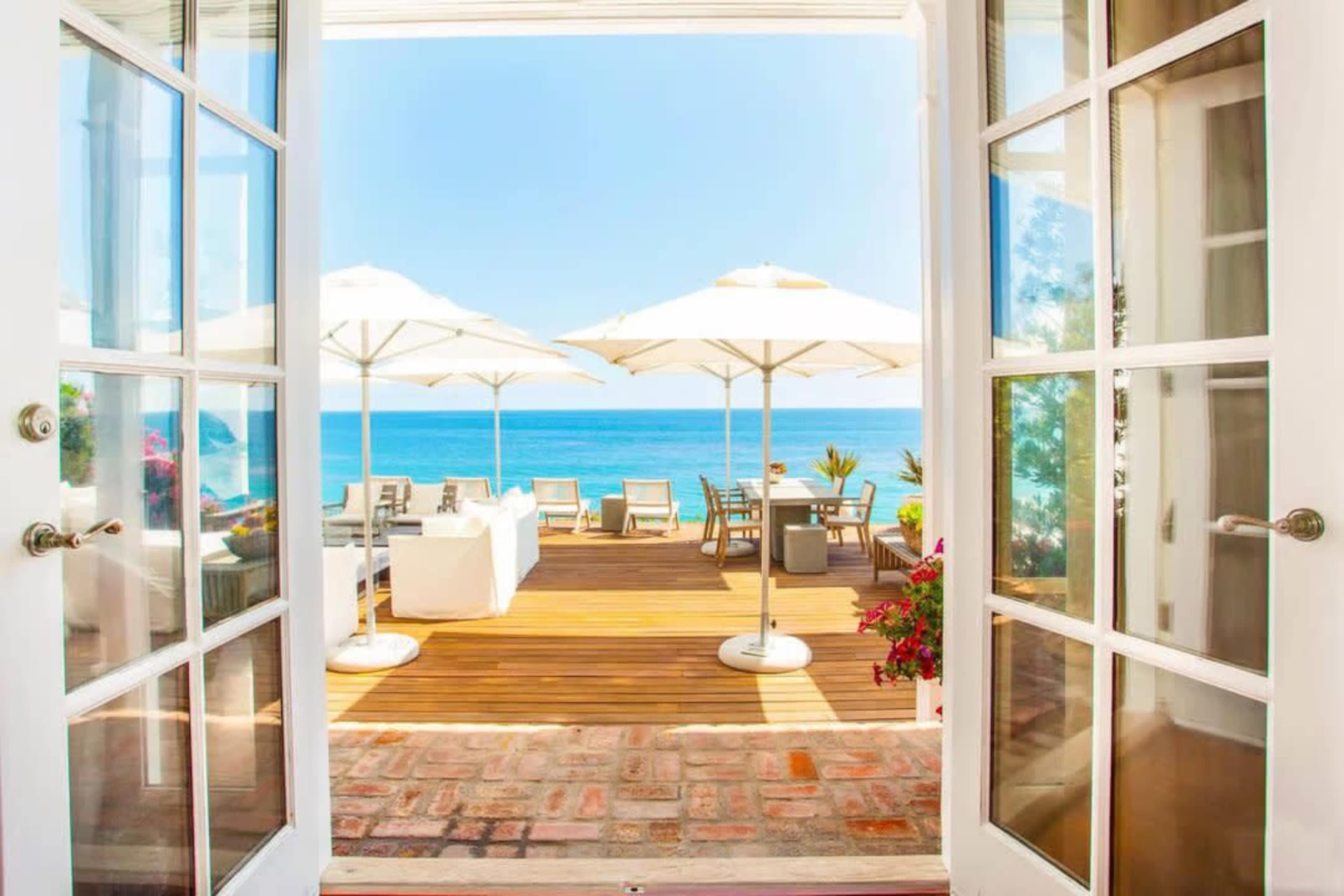 A set of double doors opens to a deck with lounge seating and umbrellas facing a calm blue sea.