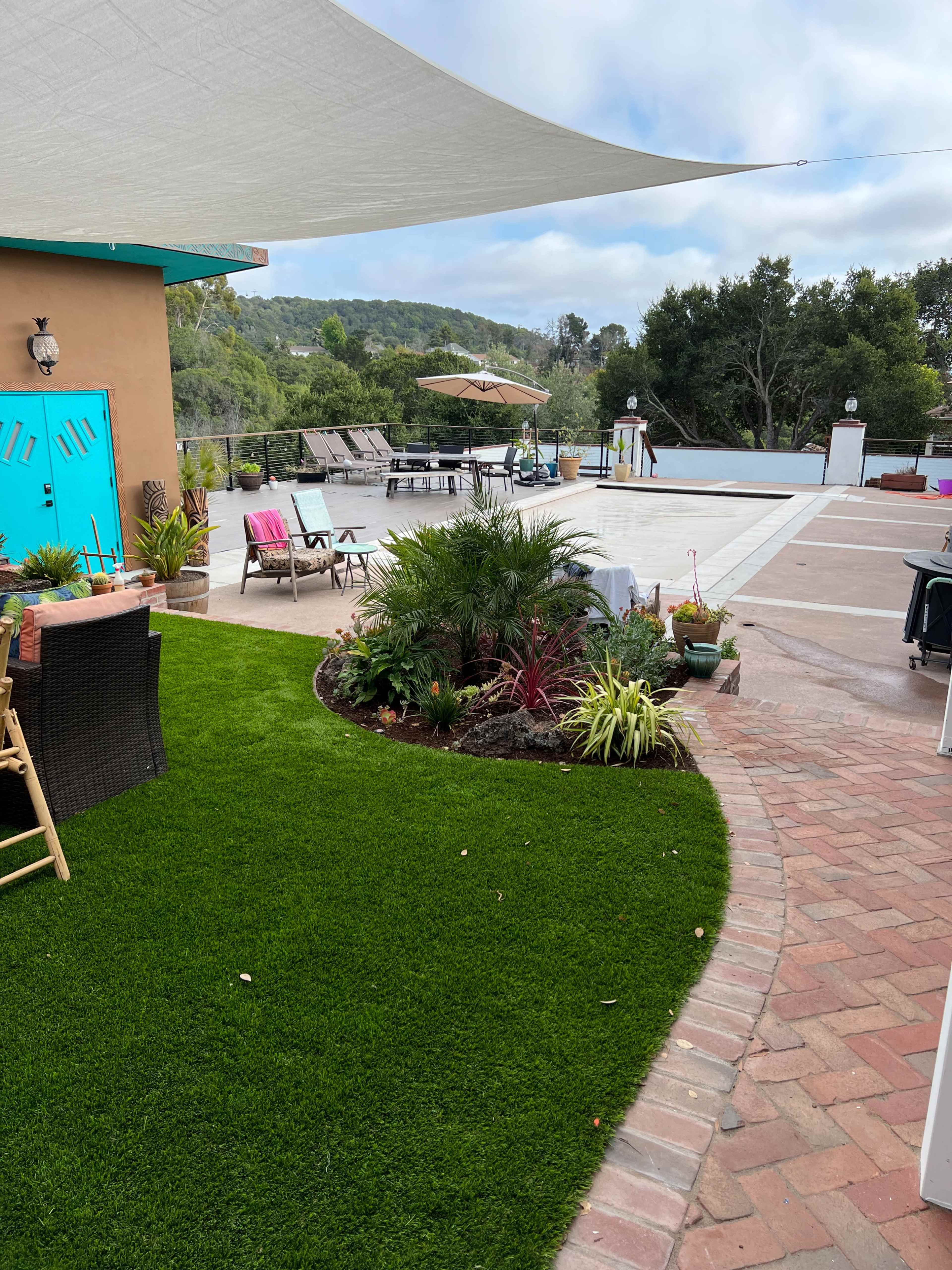 A well-landscaped outdoor area with artificial grass, a variety of plants, and a swimming pool in the background, surrounded by lounge chairs and umbrellas.