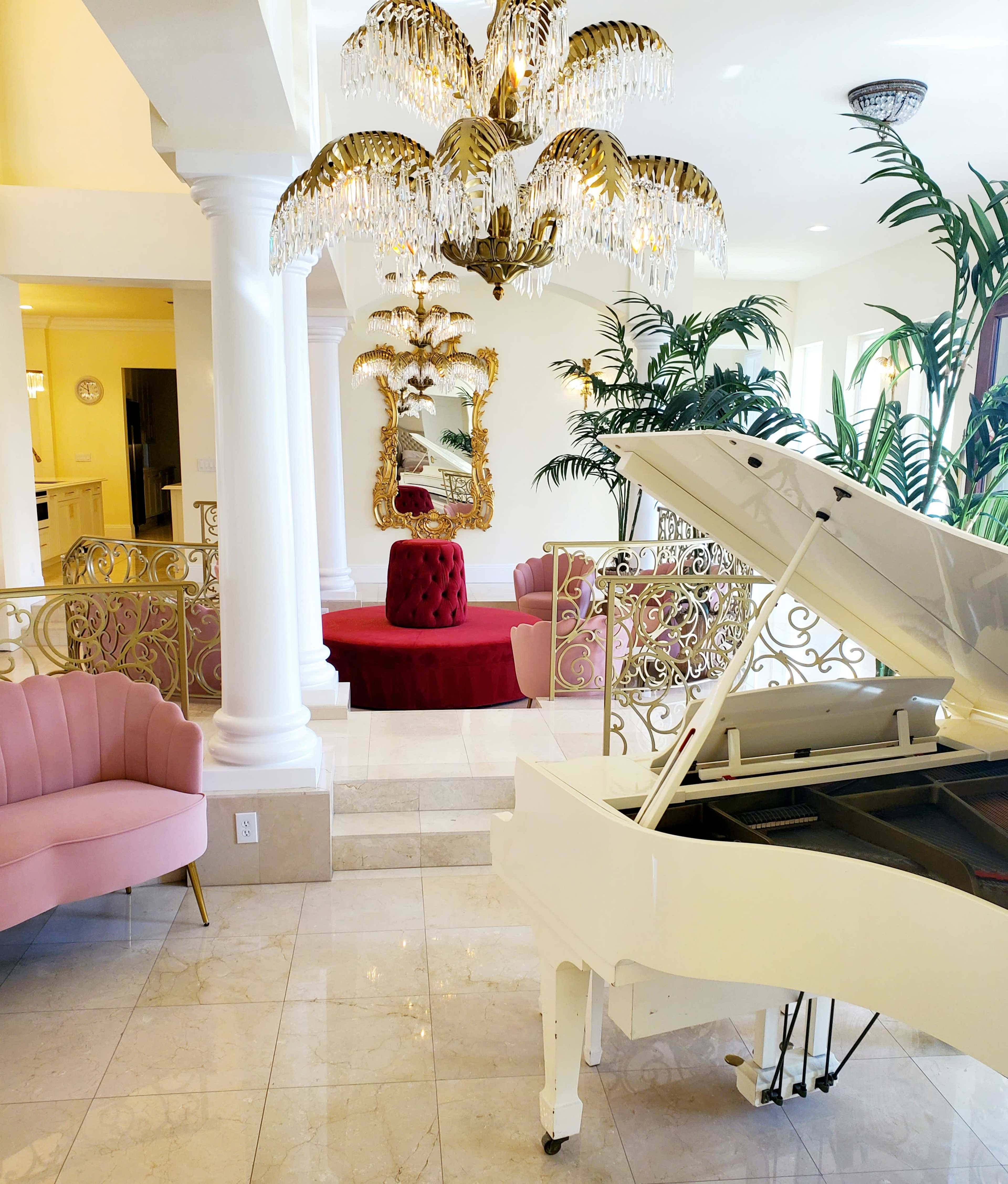 The image depicts a brightly lit room featuring a grand white piano, pink and red upholstered seating, tall indoor plants, and an ornate chandelier.