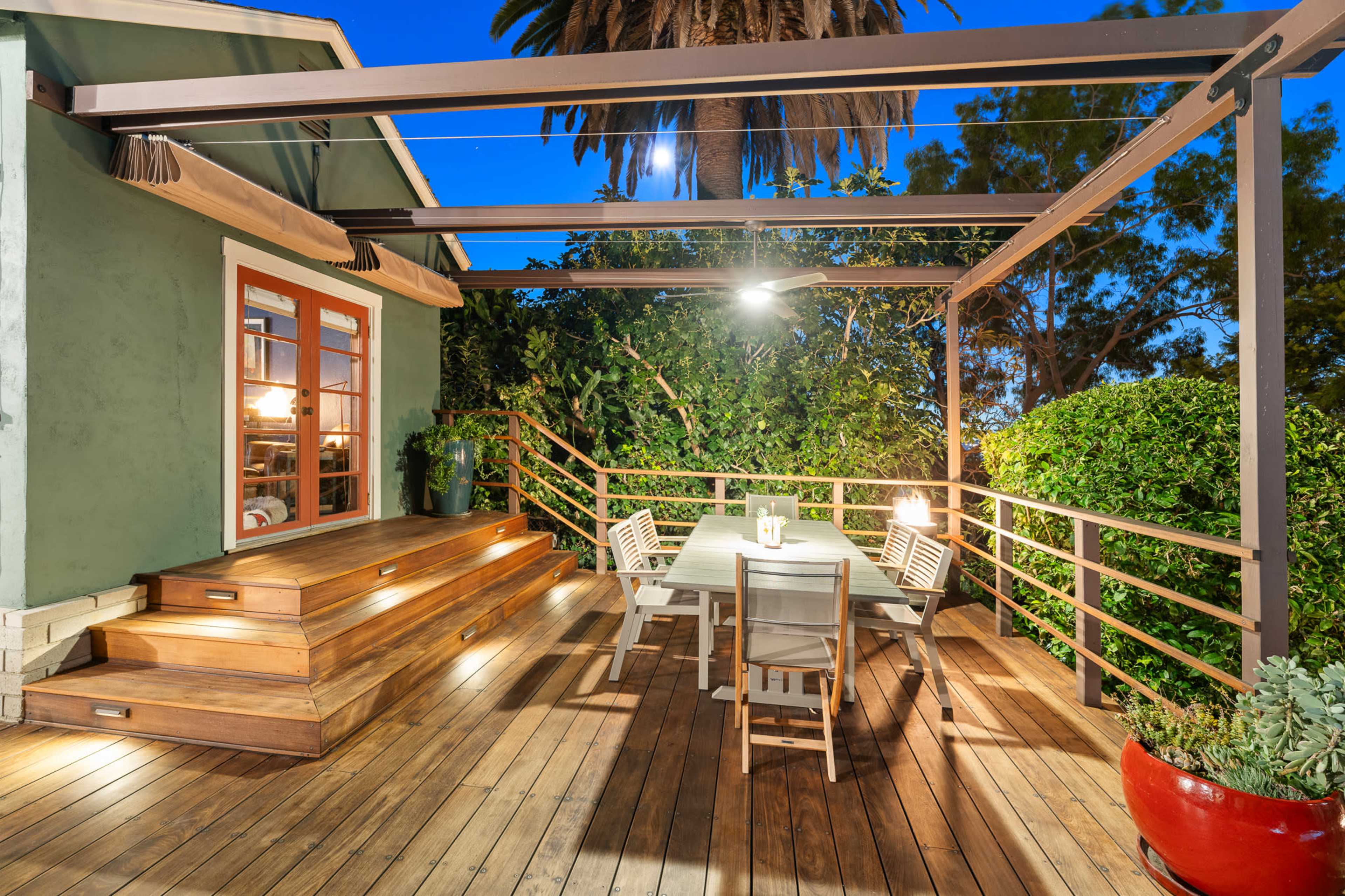 The image shows a wooden deck with a dining table and chairs, surrounded by greenery and illuminated by lights under a pergola.