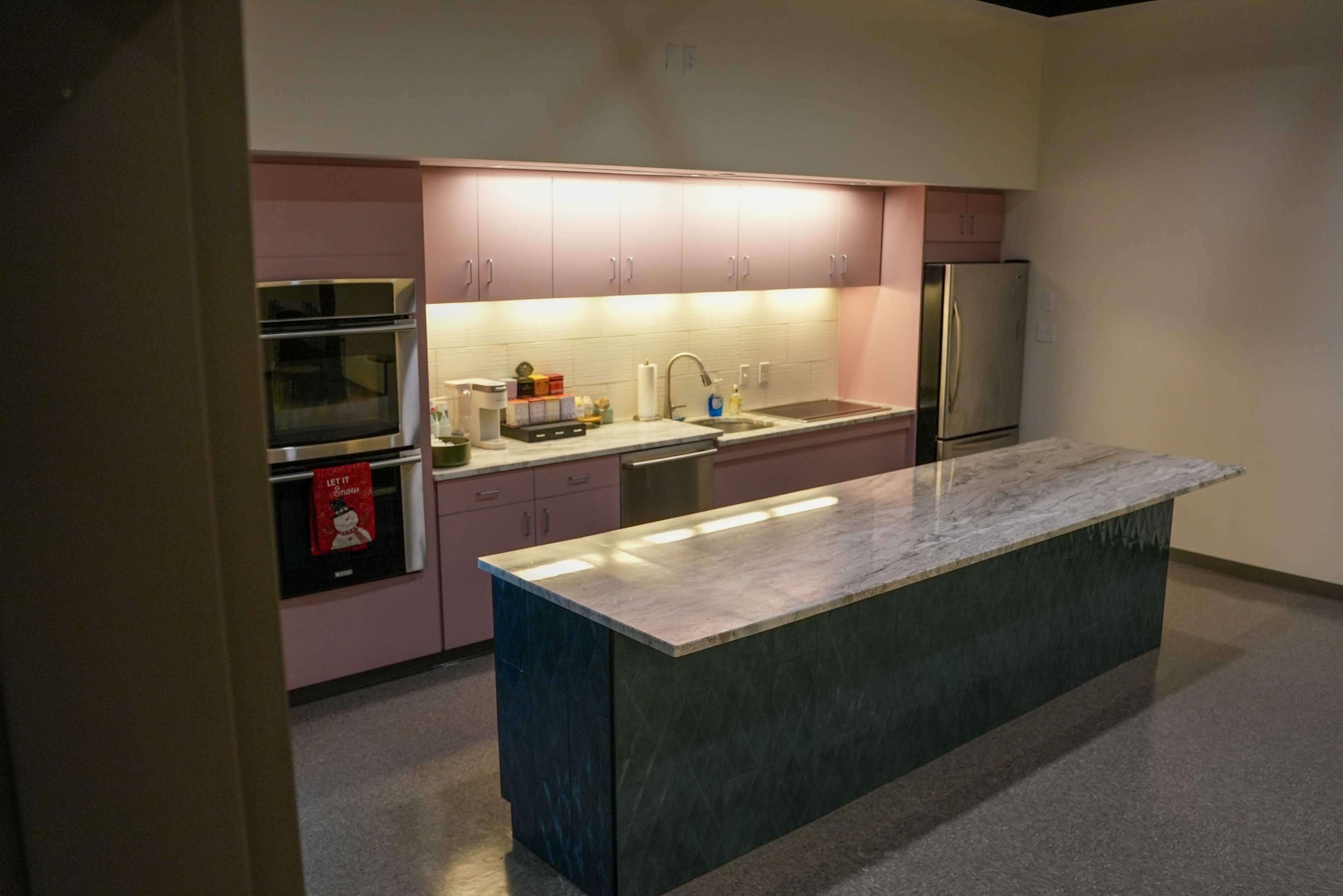 The image shows a contemporary kitchen featuring pink cabinetry, a large marble countertop island, and stainless steel appliances.