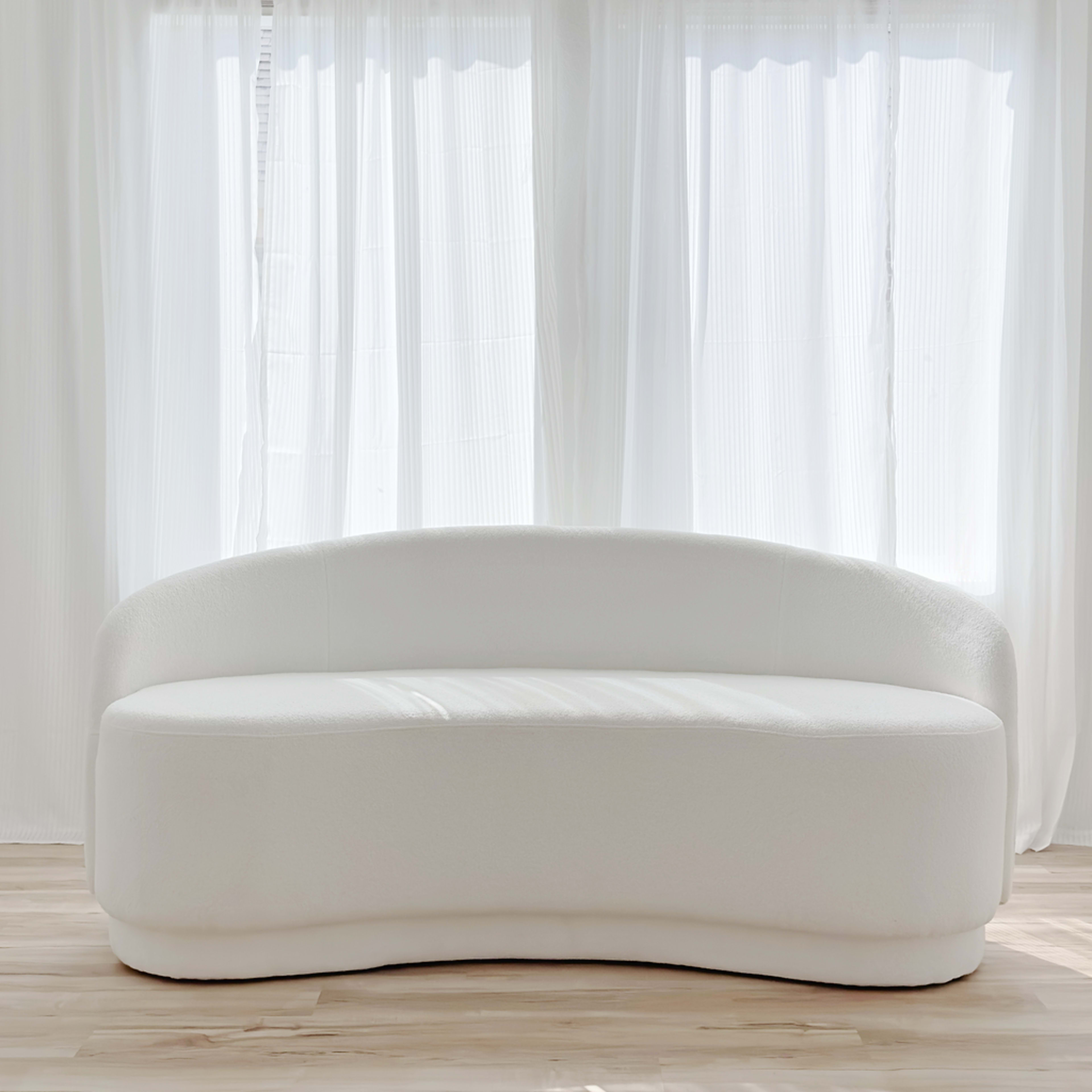 A curved, white sofa is positioned in front of large windows covered with sheer curtains.