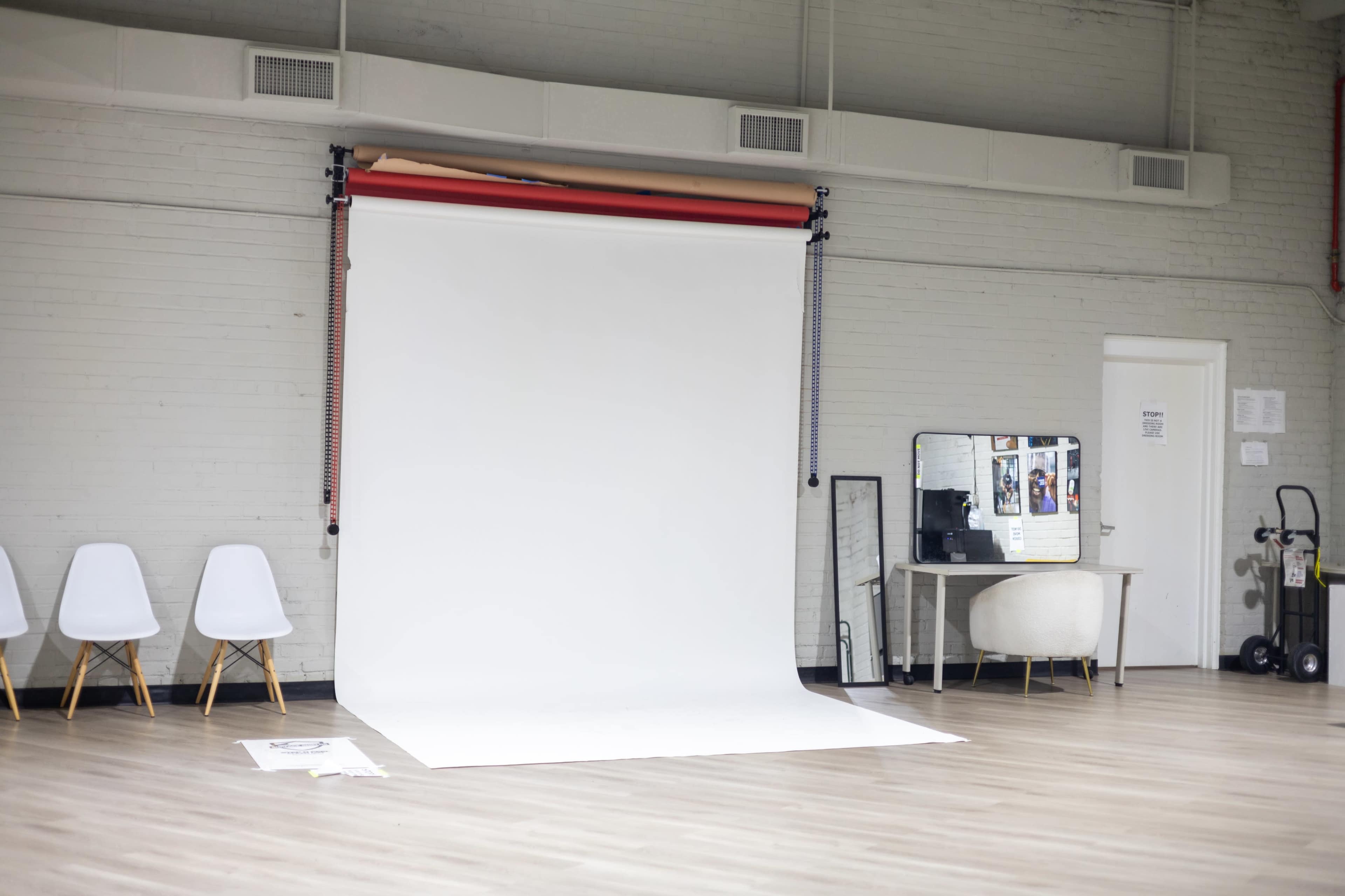 The image shows an empty photography studio with a white backdrop, a small desk with a mirror, and a few white chairs.