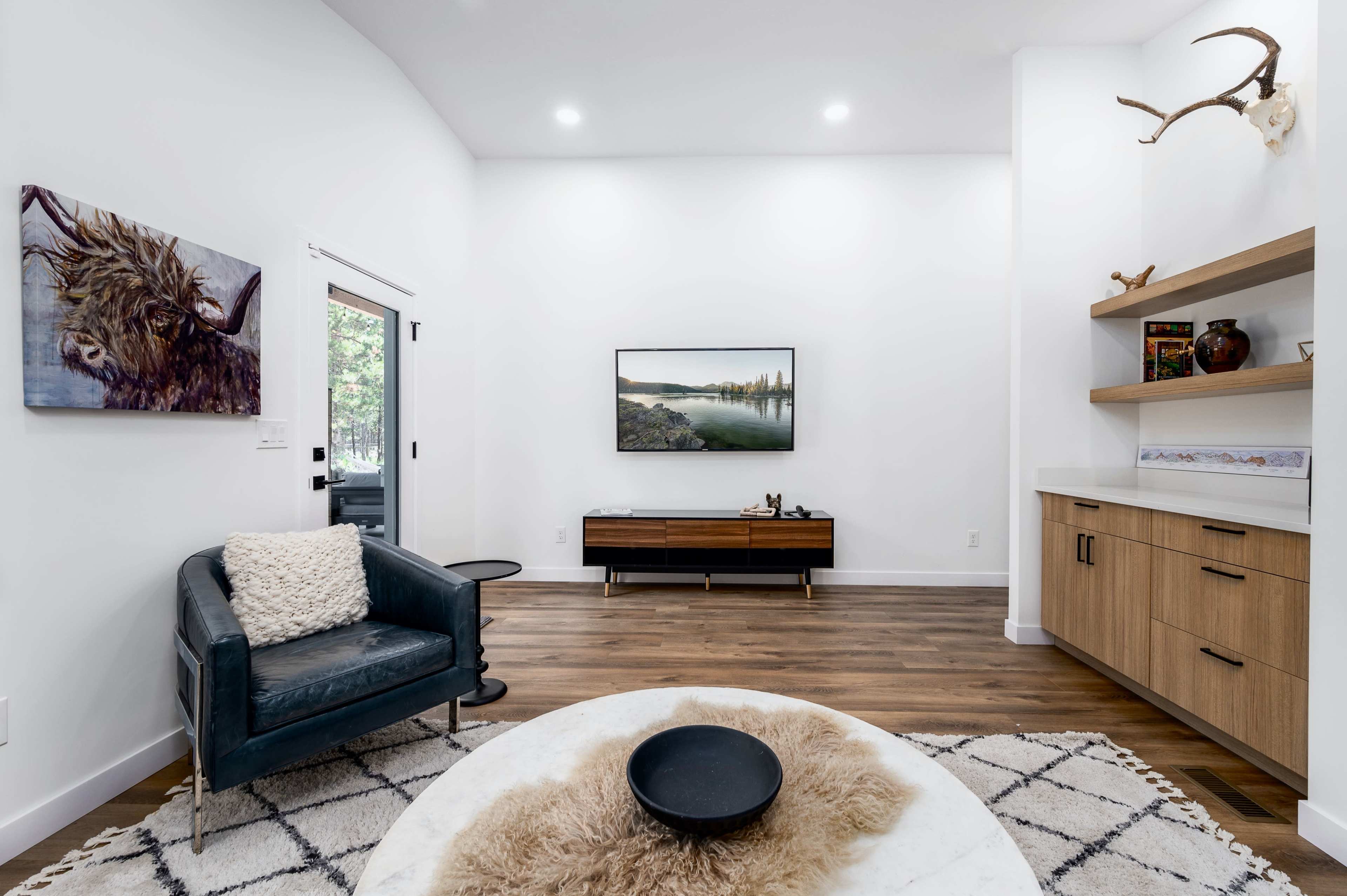 A modern living room featuring a television mounted on a white wall, a wooden media console, a single armchair, and a round coffee table with a decorative item on it.