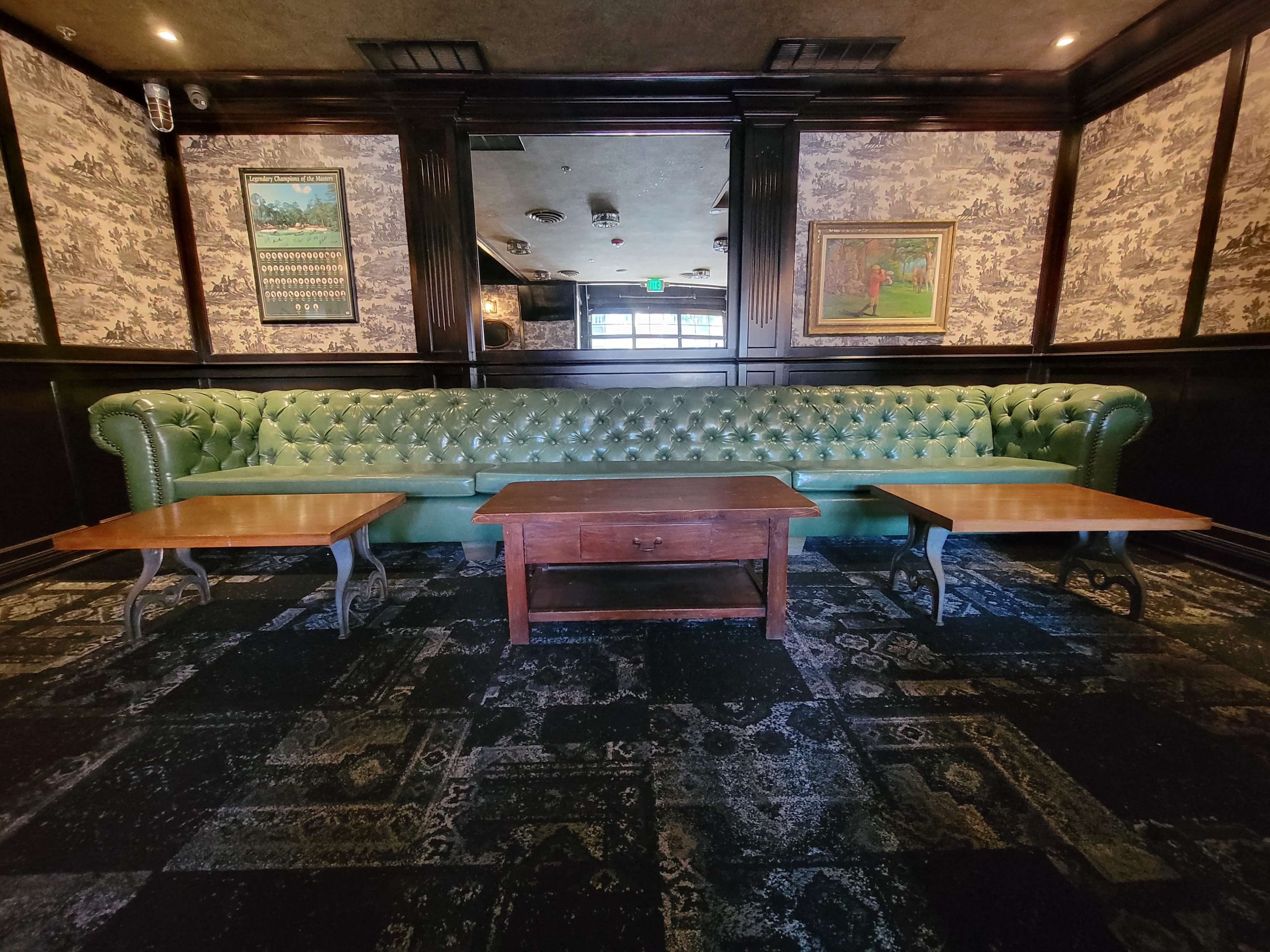 A darkly decorated interior space with green leather seating, wooden tables, and framed artwork on the walls.