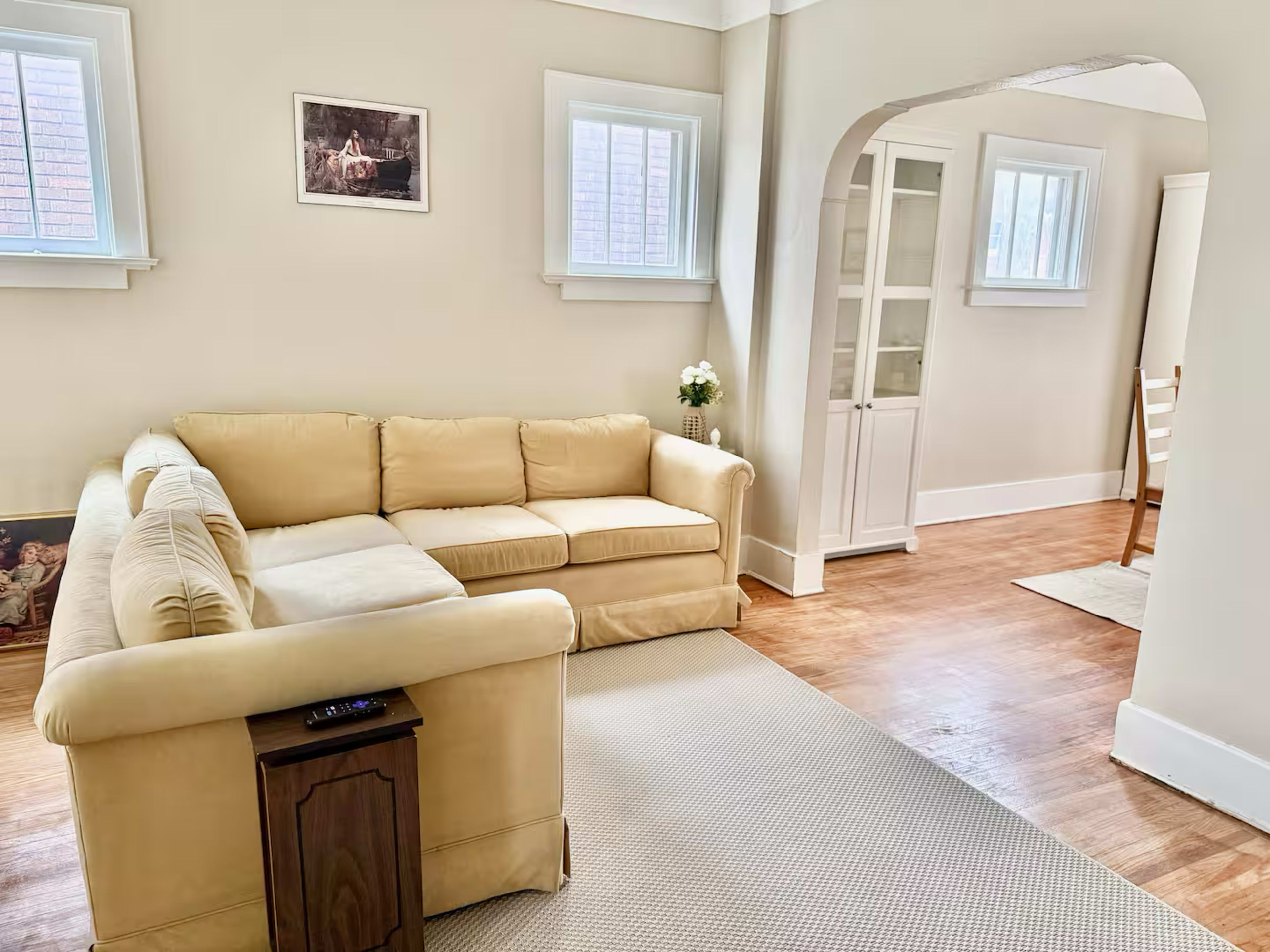 The image shows a light-colored living room with a beige sectional couch and a dining area visible in the background.