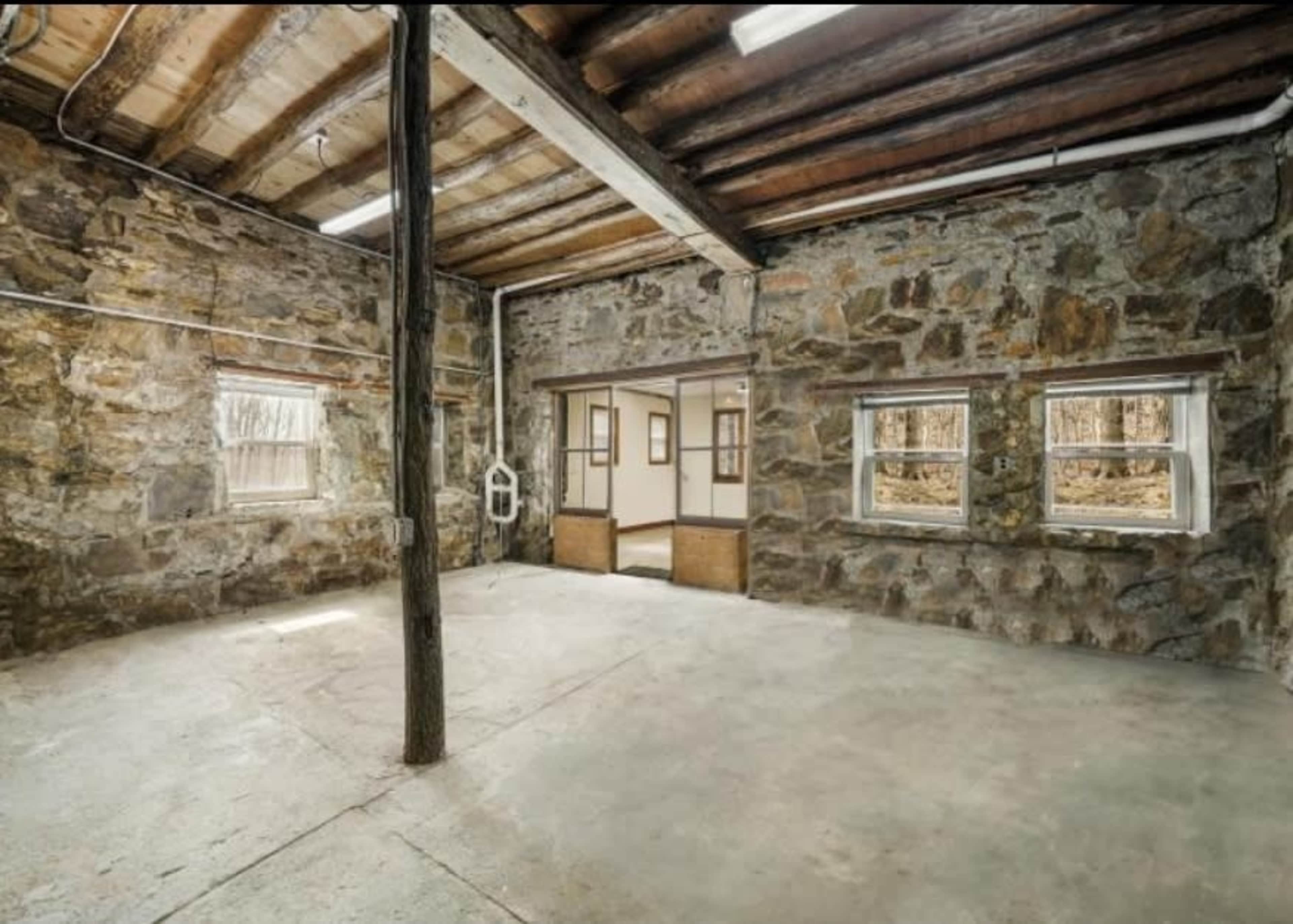 The image shows an interior space with stone walls, a concrete floor, wooden ceiling beams, and a central support pillar.