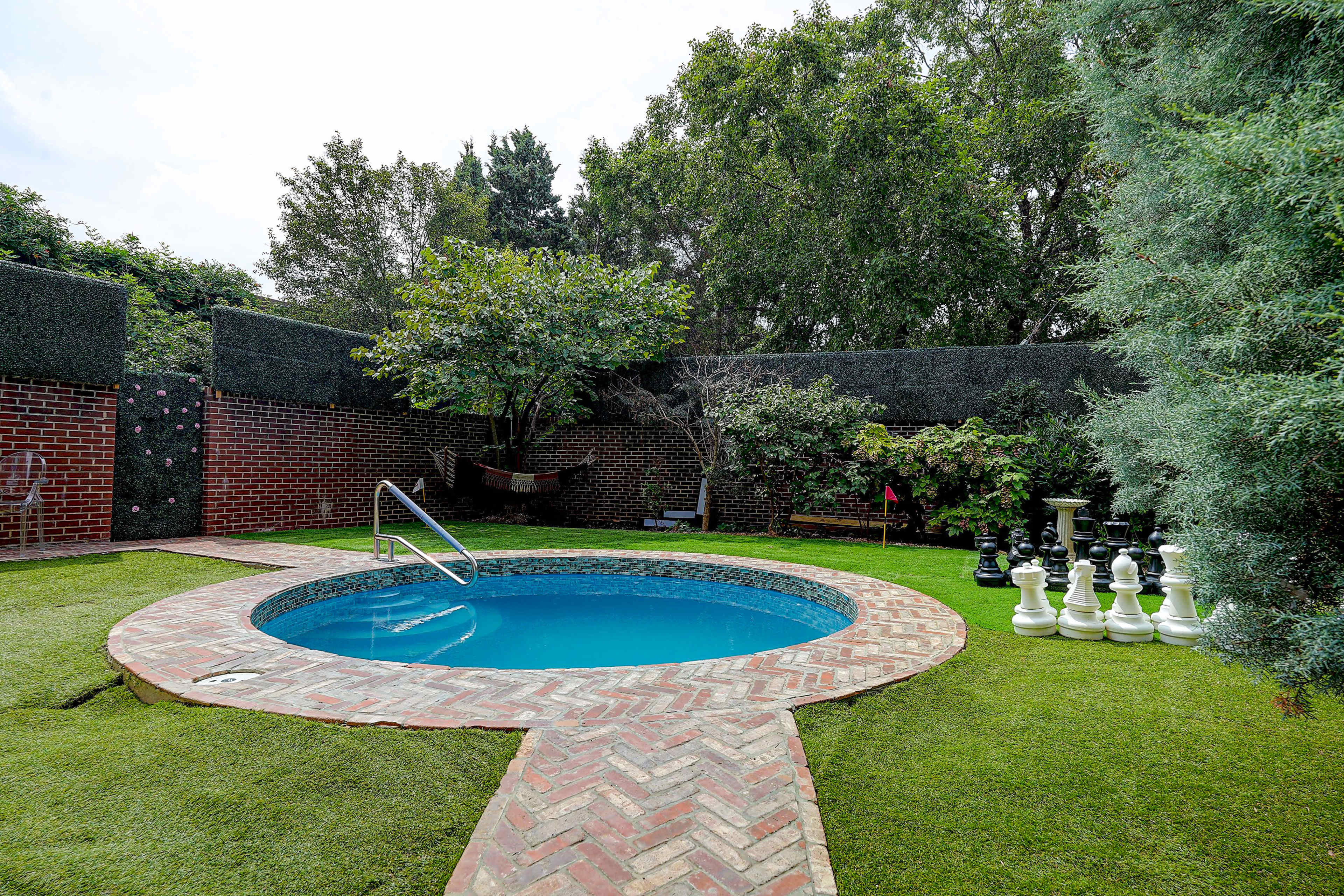A circular blue swimming pool surrounded by a brick pathway, lush greenery, and a large chess set on a grassy area.