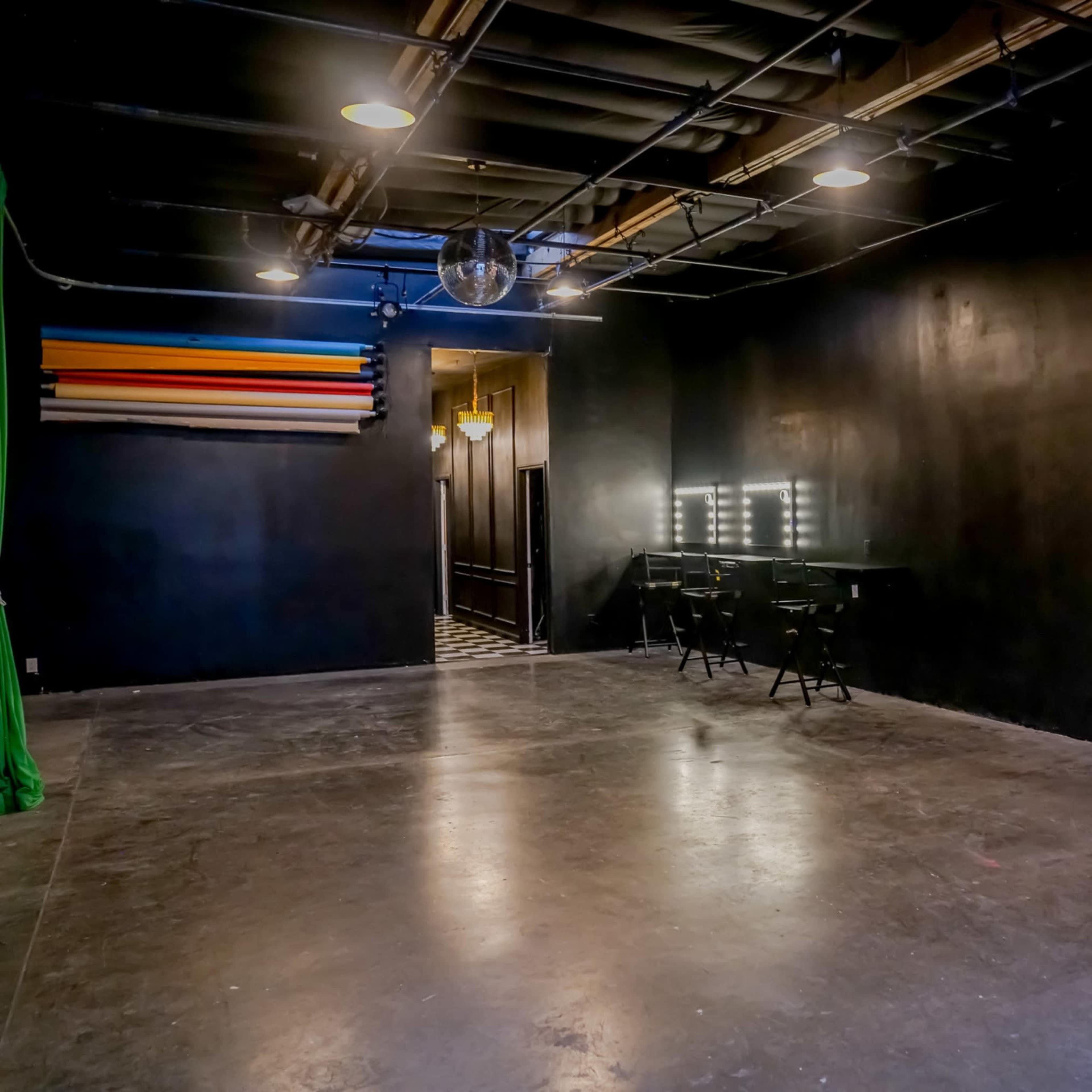 The image shows a spacious, dimly lit studio with black walls, a reflective disco ball, a doorway leading to another room, and a vanity area with mirrors and chairs.