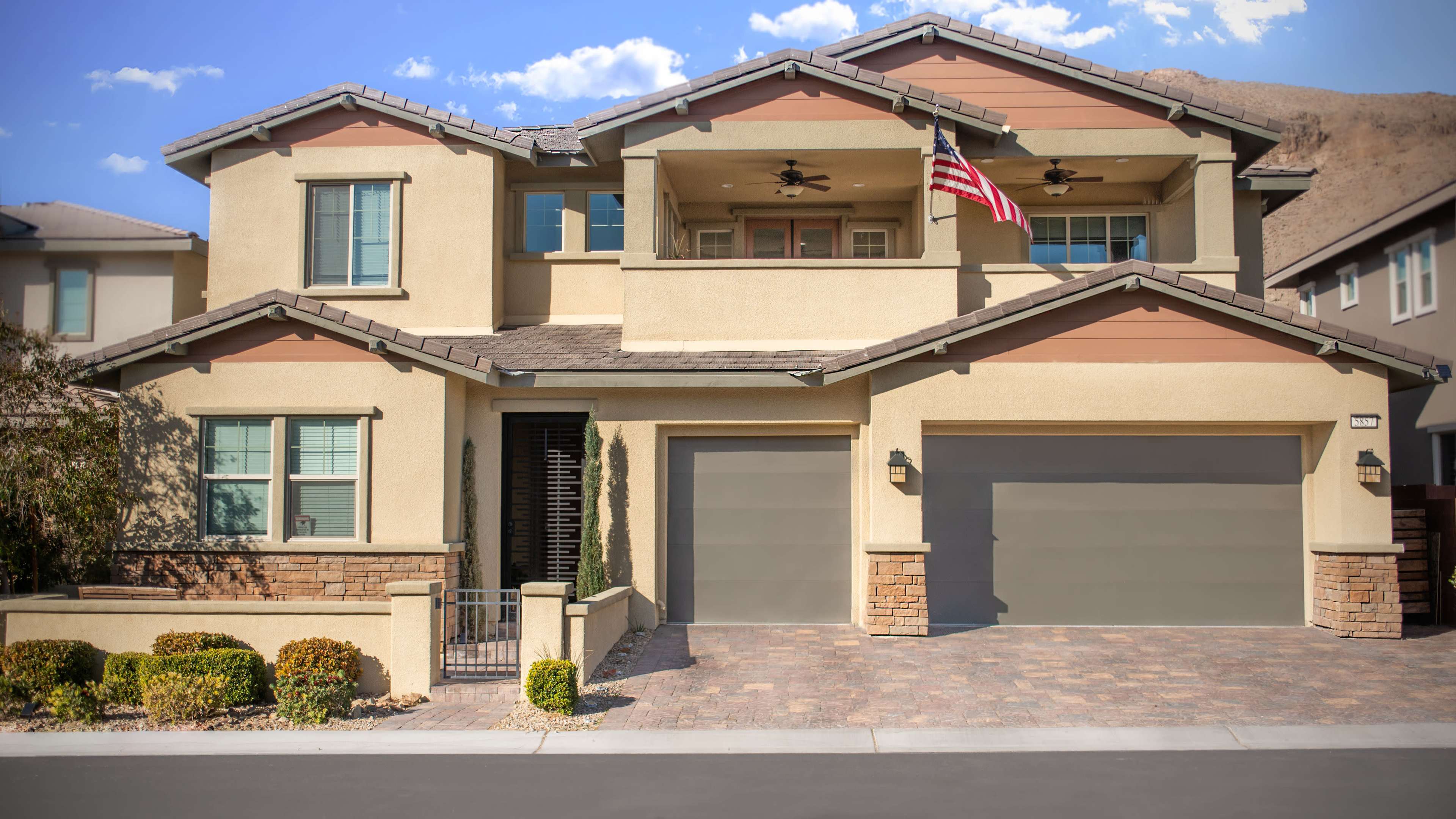 A two-story house with a stucco exterior features a front porch, two garage doors, and an American flag displayed on the balcony.