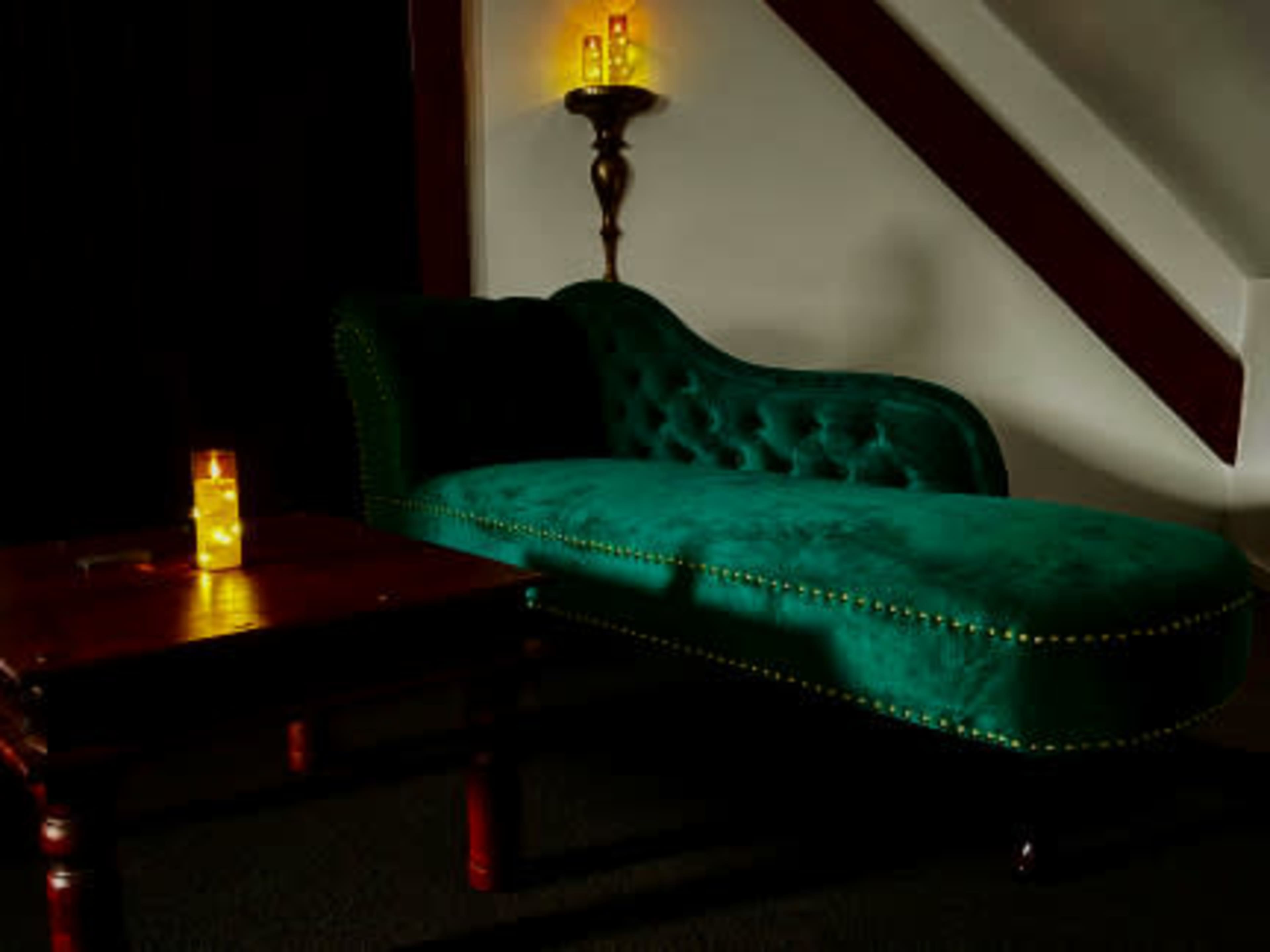 A green velvet chaise lounge is positioned beside a wooden table and is illuminated by candlelight in a dimly lit room.
