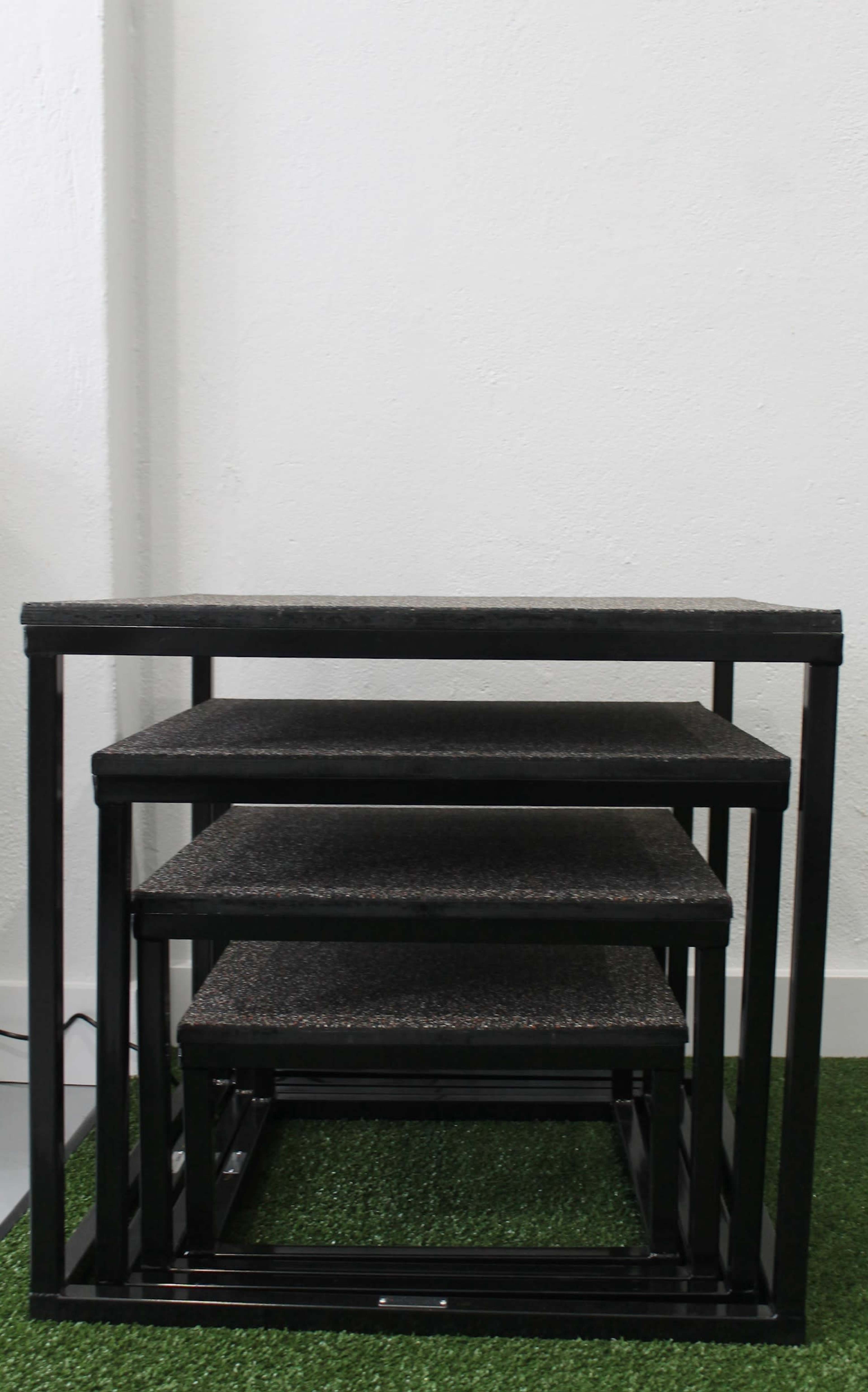 A set of four nesting tables with black frames and textured surfaces, arranged in a staggered manner against a white wall.