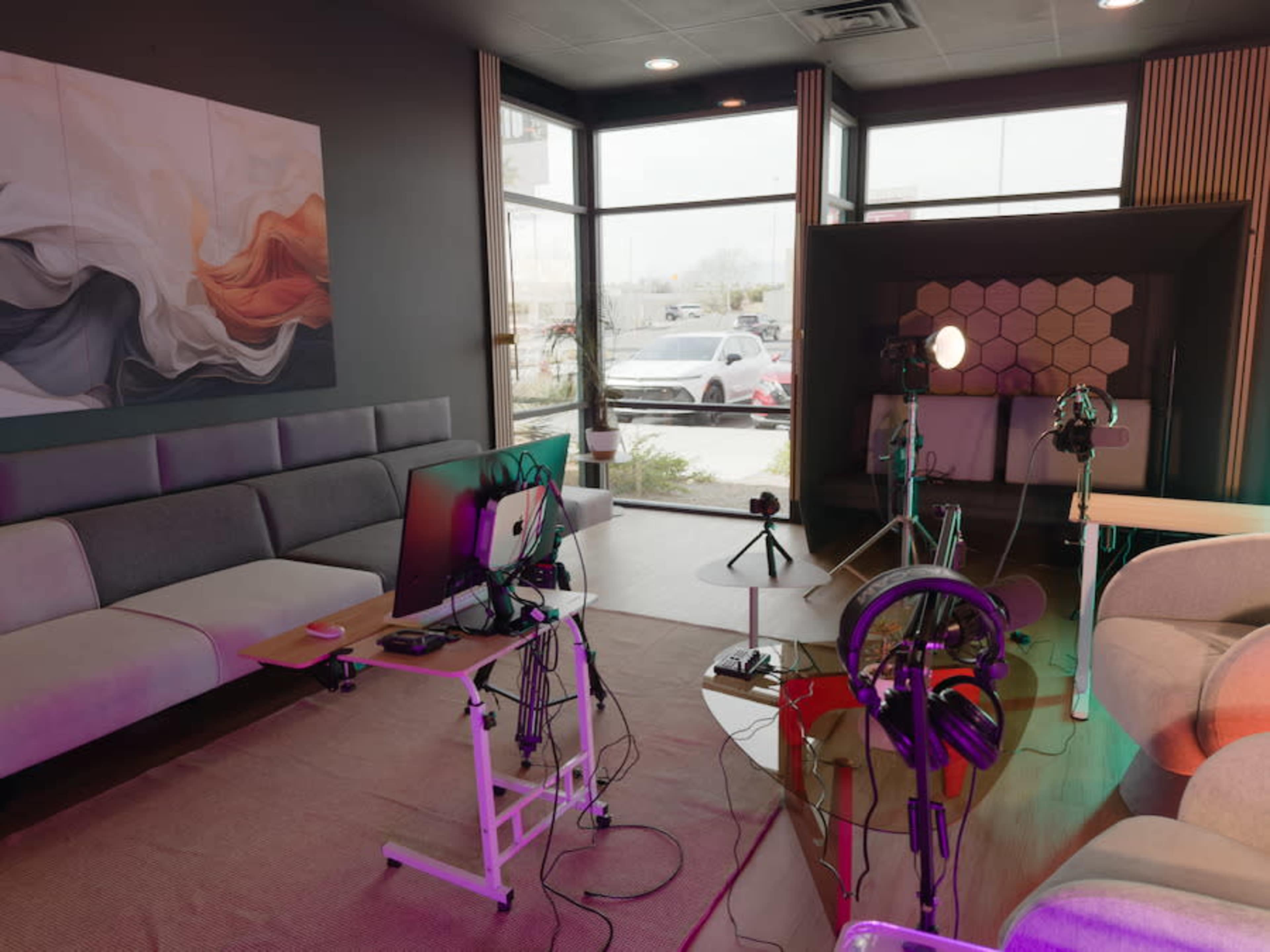 The image shows a modern studio space featuring a gray sofa, a workstation with a computer and multiple cables, and lighting equipment set up for a recording session.