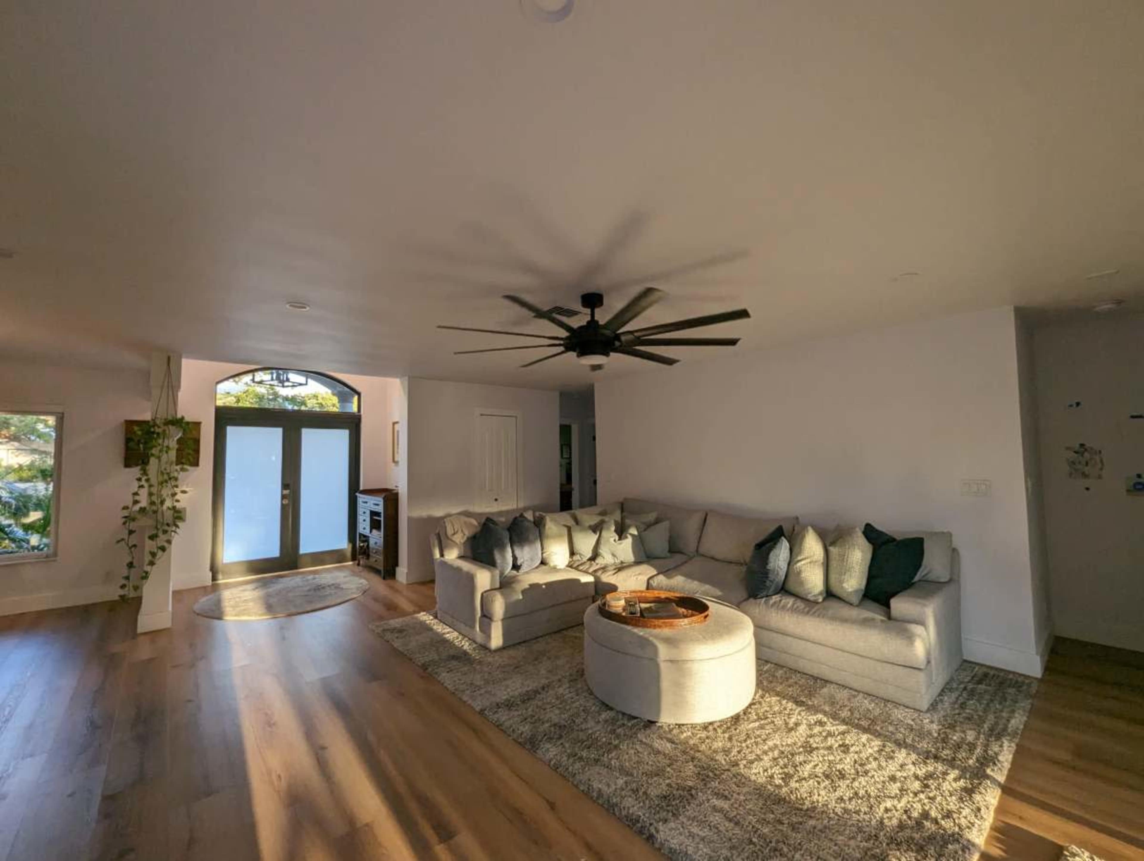 A spacious living room features a light-colored sectional sofa, a circular coffee table, and a large ceiling fan, with natural light streaming in through a large front door.