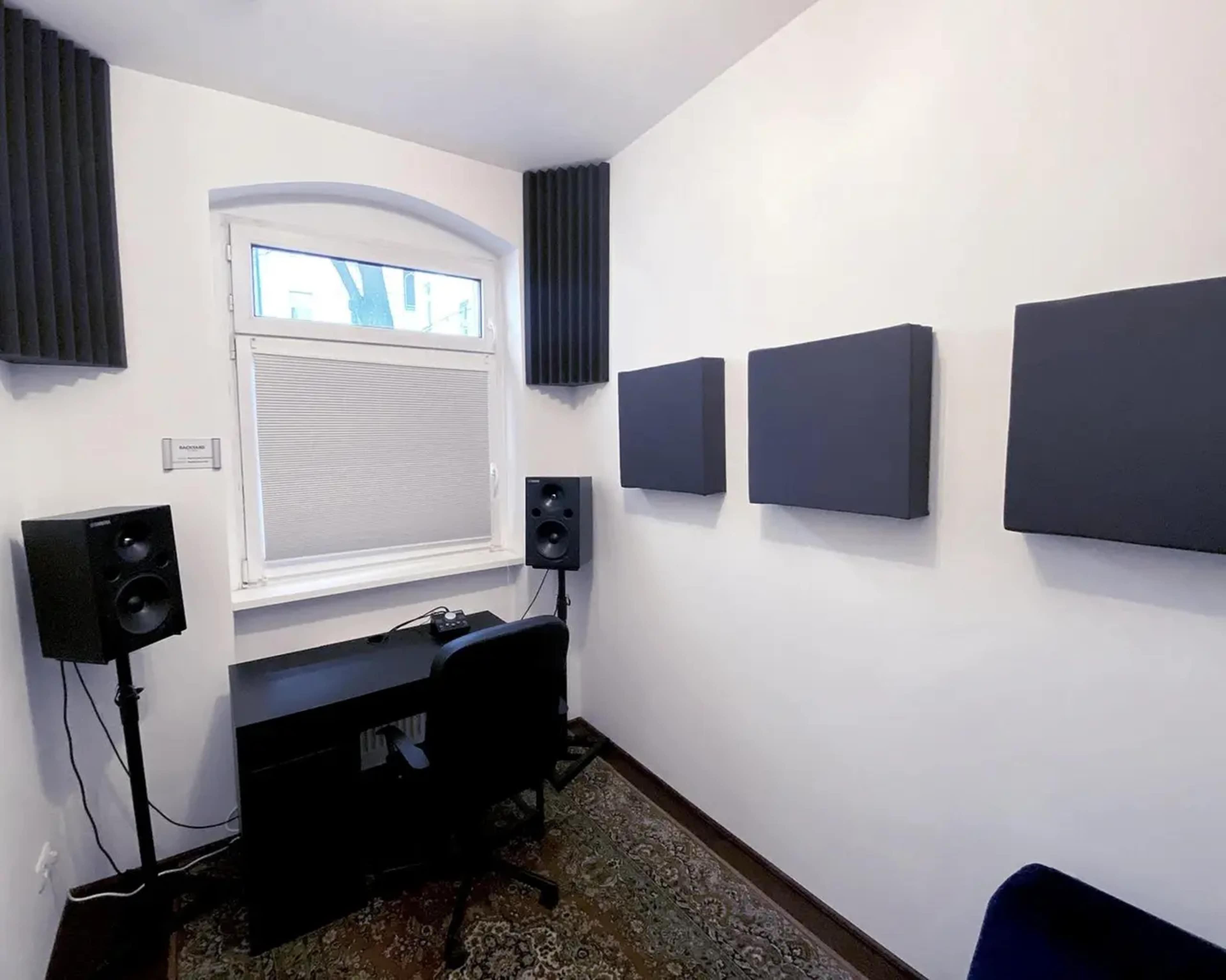 A small audio recording studio with soundproofing panels on the walls, a desk, and speakers positioned near a window.