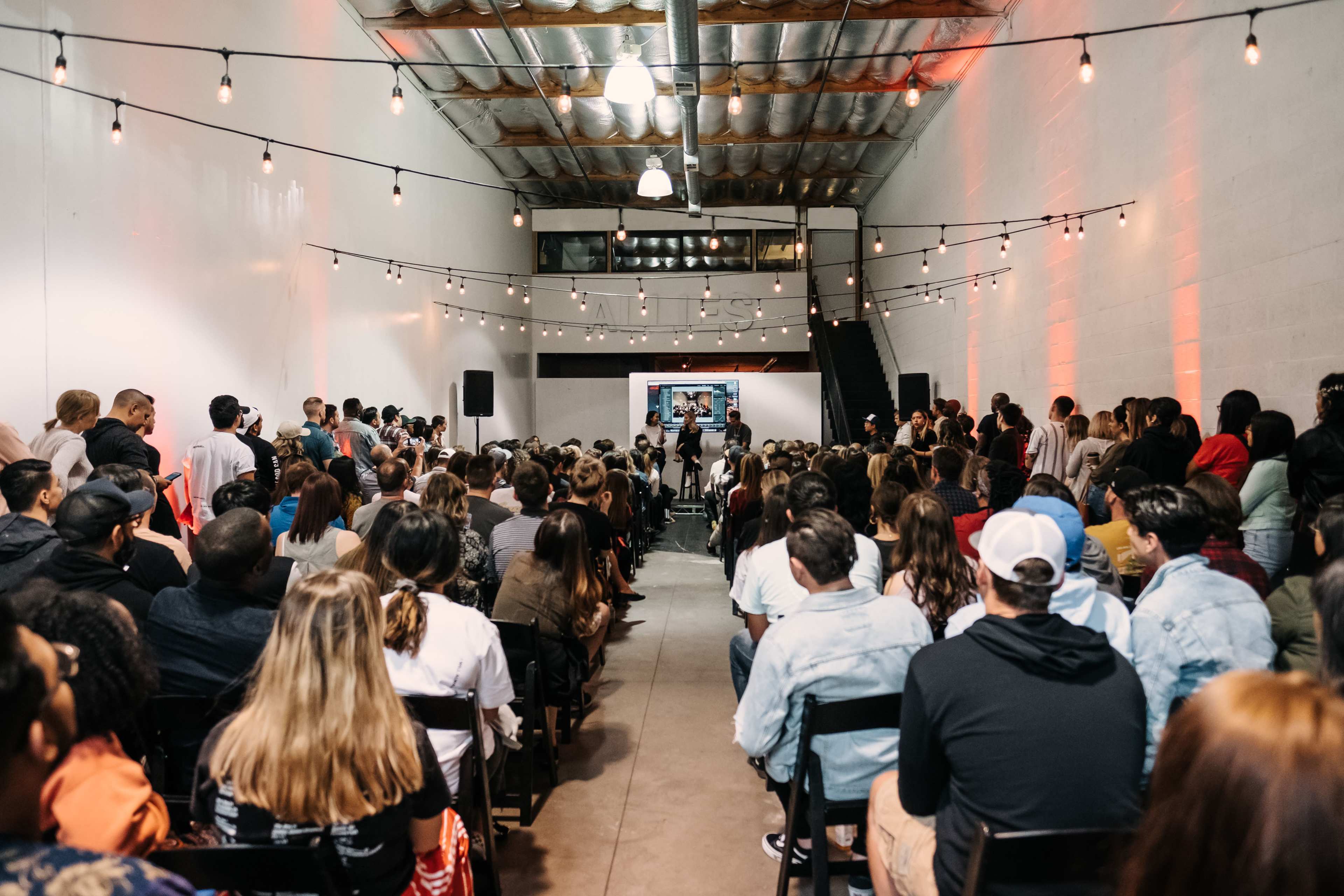 A large audience is seated in a brightly lit warehouse space, facing a stage where a speaker addresses them, with string lights hanging from the ceiling above.