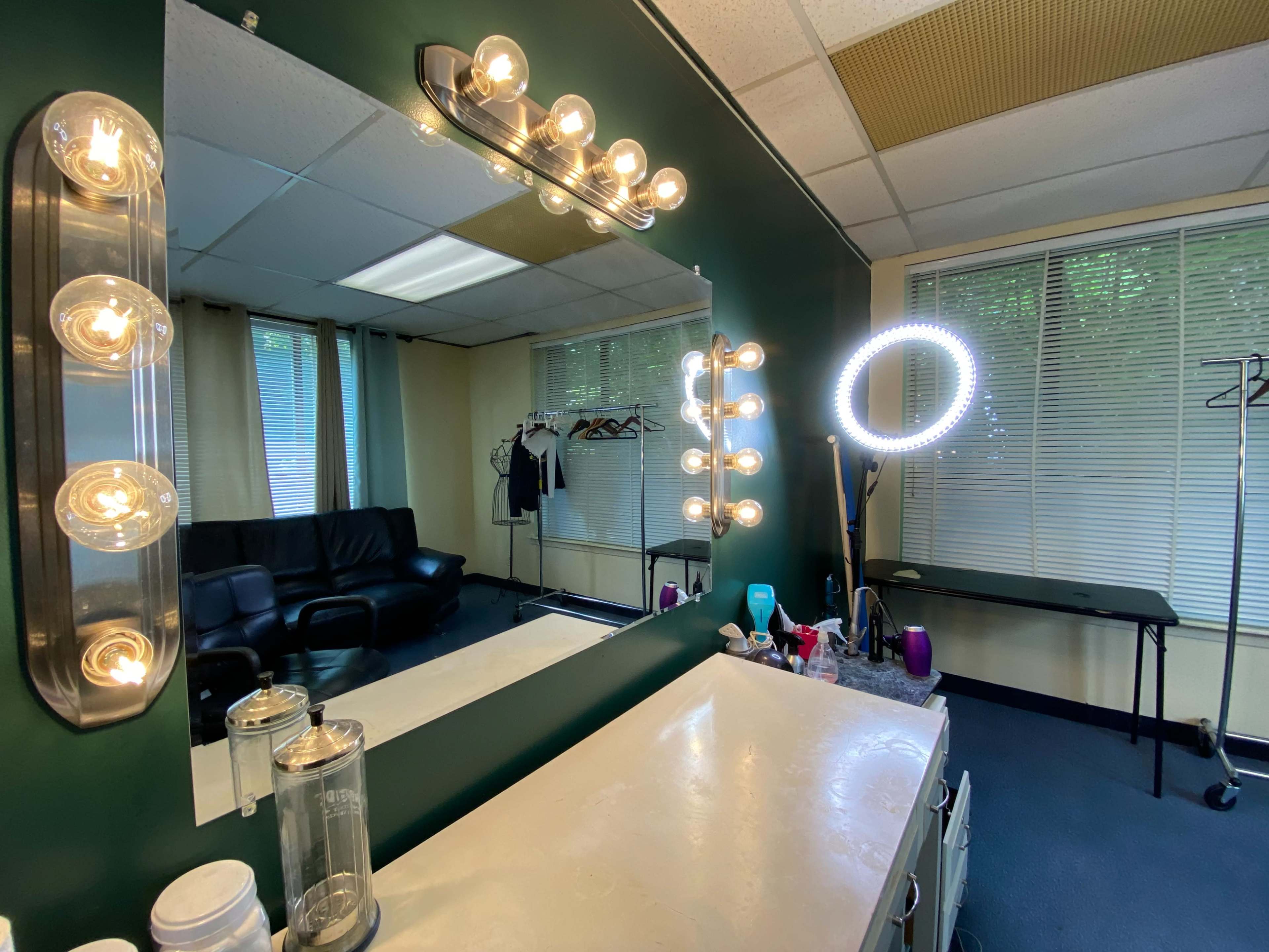 A vanity area is reflected in a large mirror, surrounded by bright lights, with a seating area and clothing rack visible in the background.