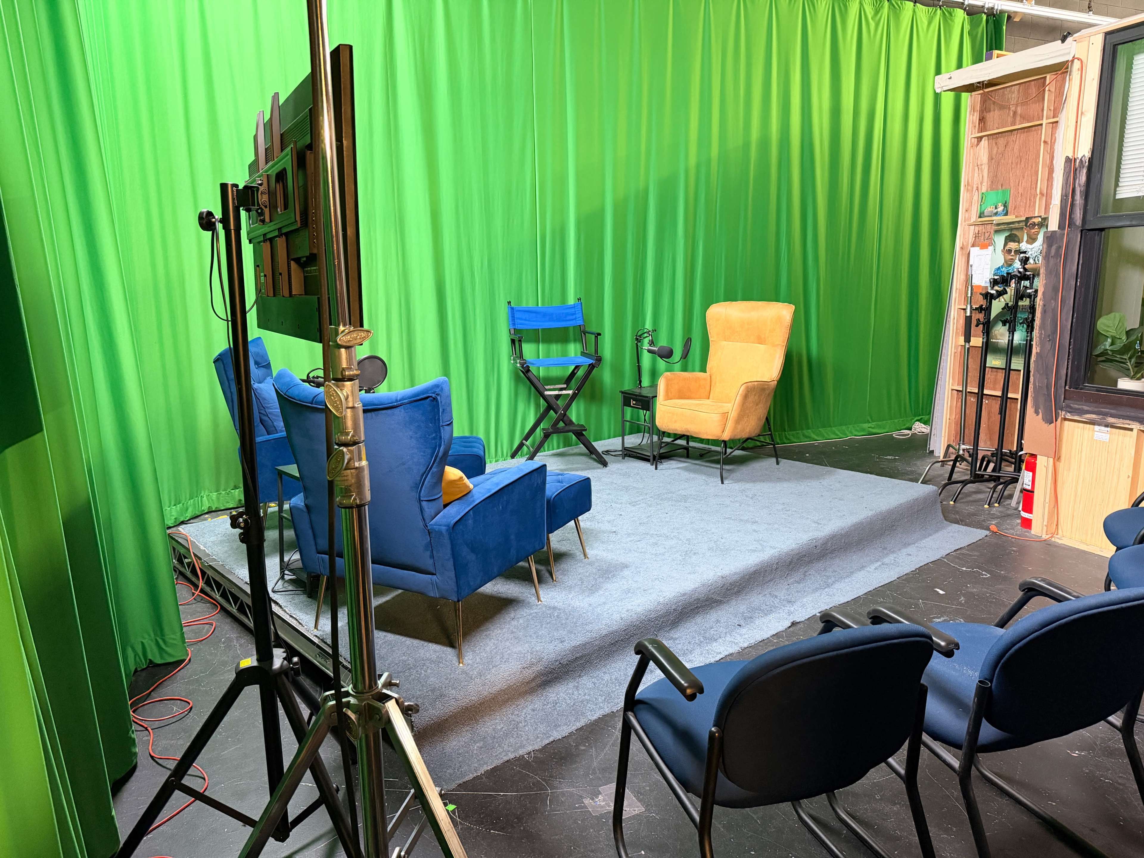 The image shows a studio setup with a green backdrop, featuring two chairs, a director's chair, and several camera equipment.