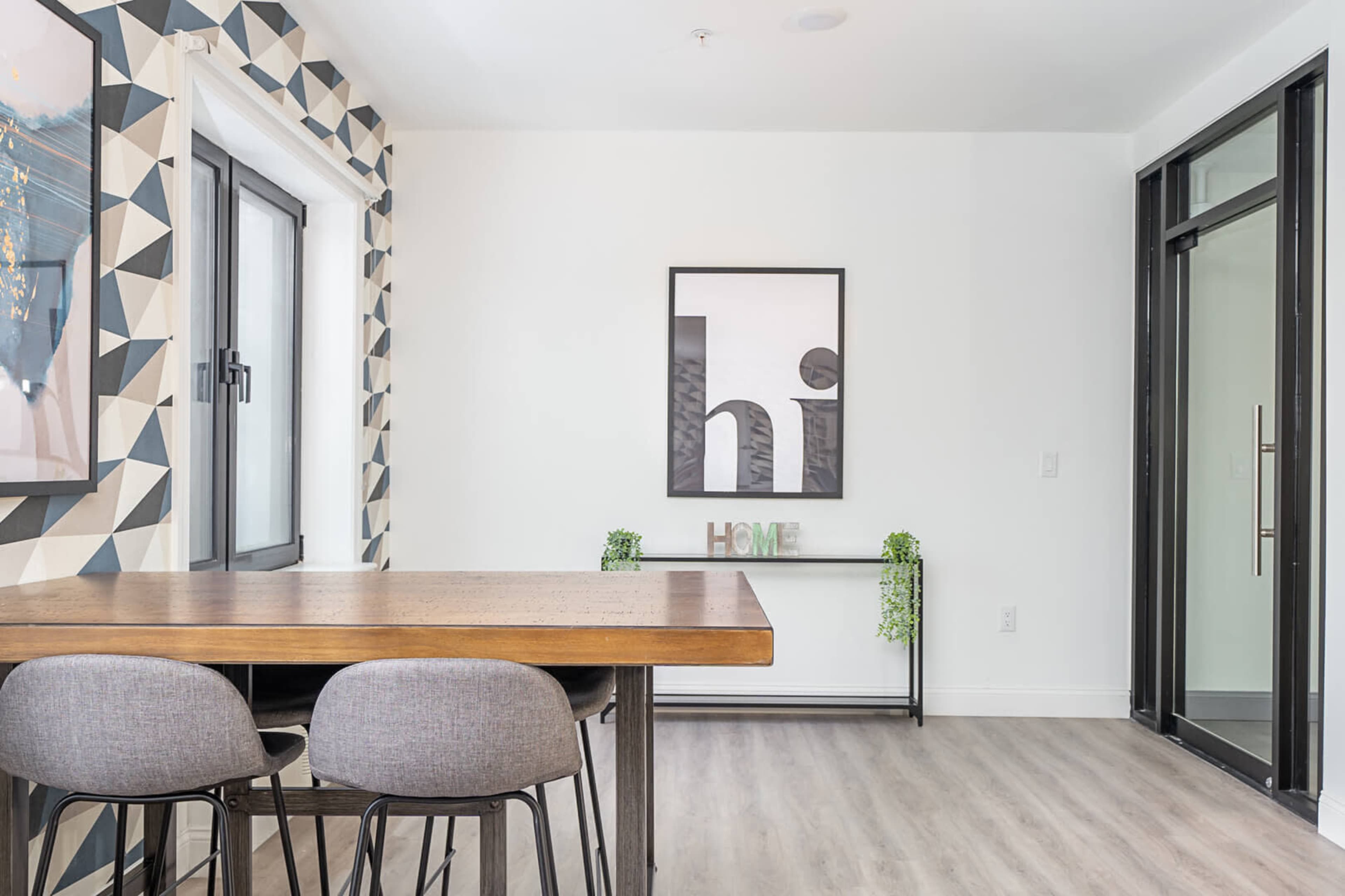 A modern dining area features a wooden table with gray chairs against a patterned wall and framed artwork.
