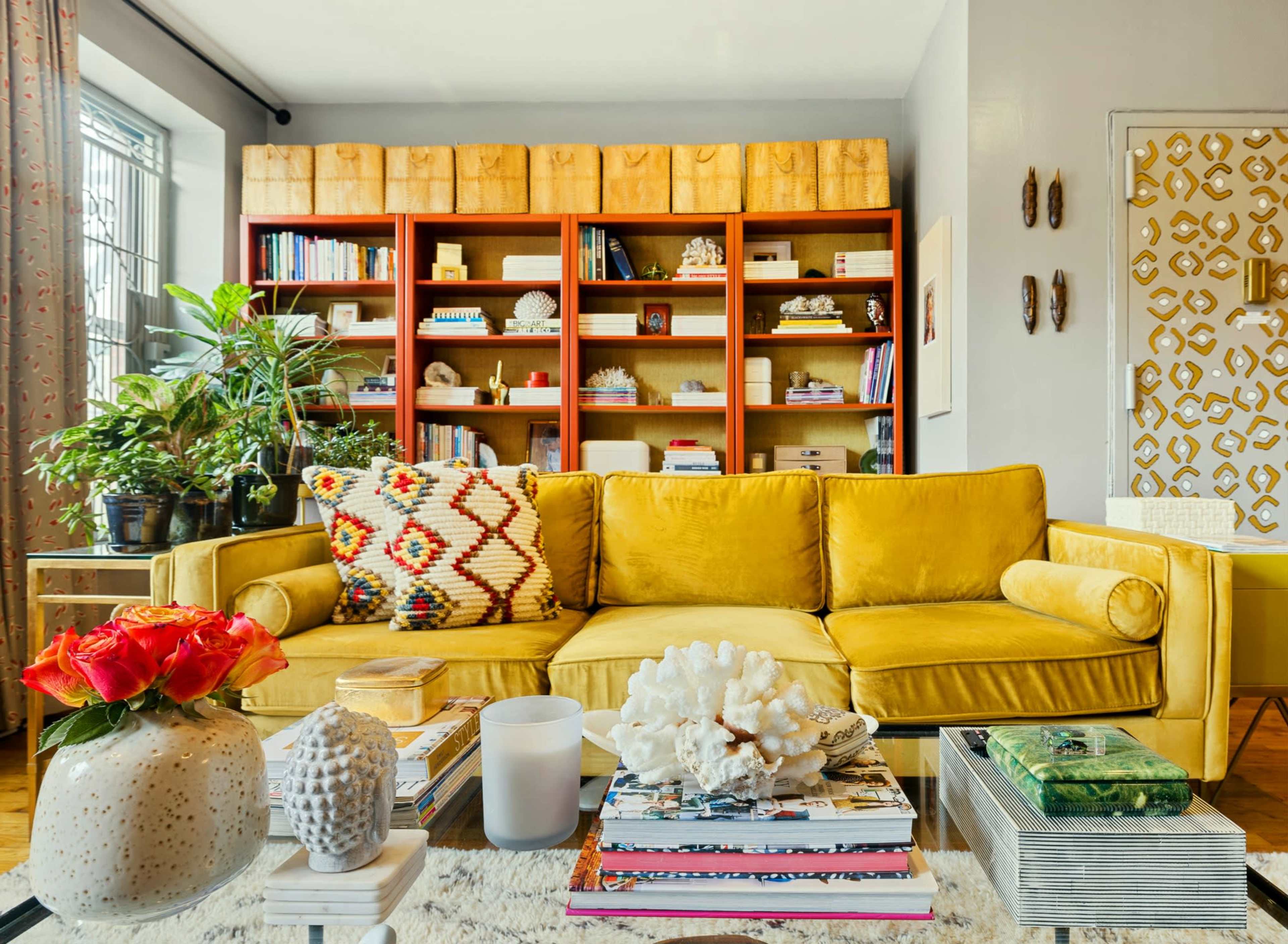 A yellow sofa with patterned pillows is positioned in front of a bookshelf filled with books and decorative items, surrounded by potted plants and a coffee table adorned with magazines and a decorative piece.