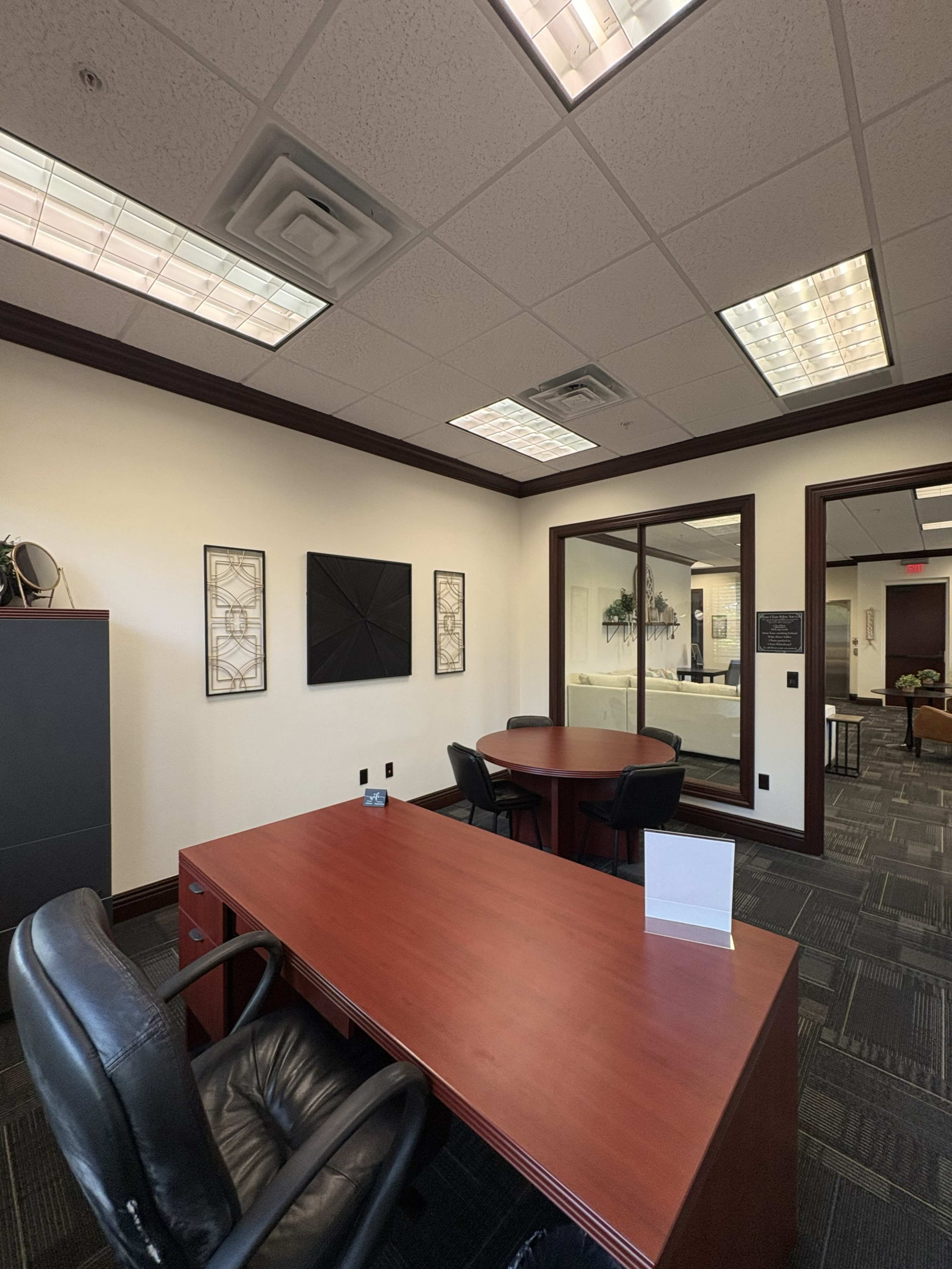 The image shows a modern office space featuring two desks with black chairs, framed wall art, and a glass-walled meeting room visible in the background.