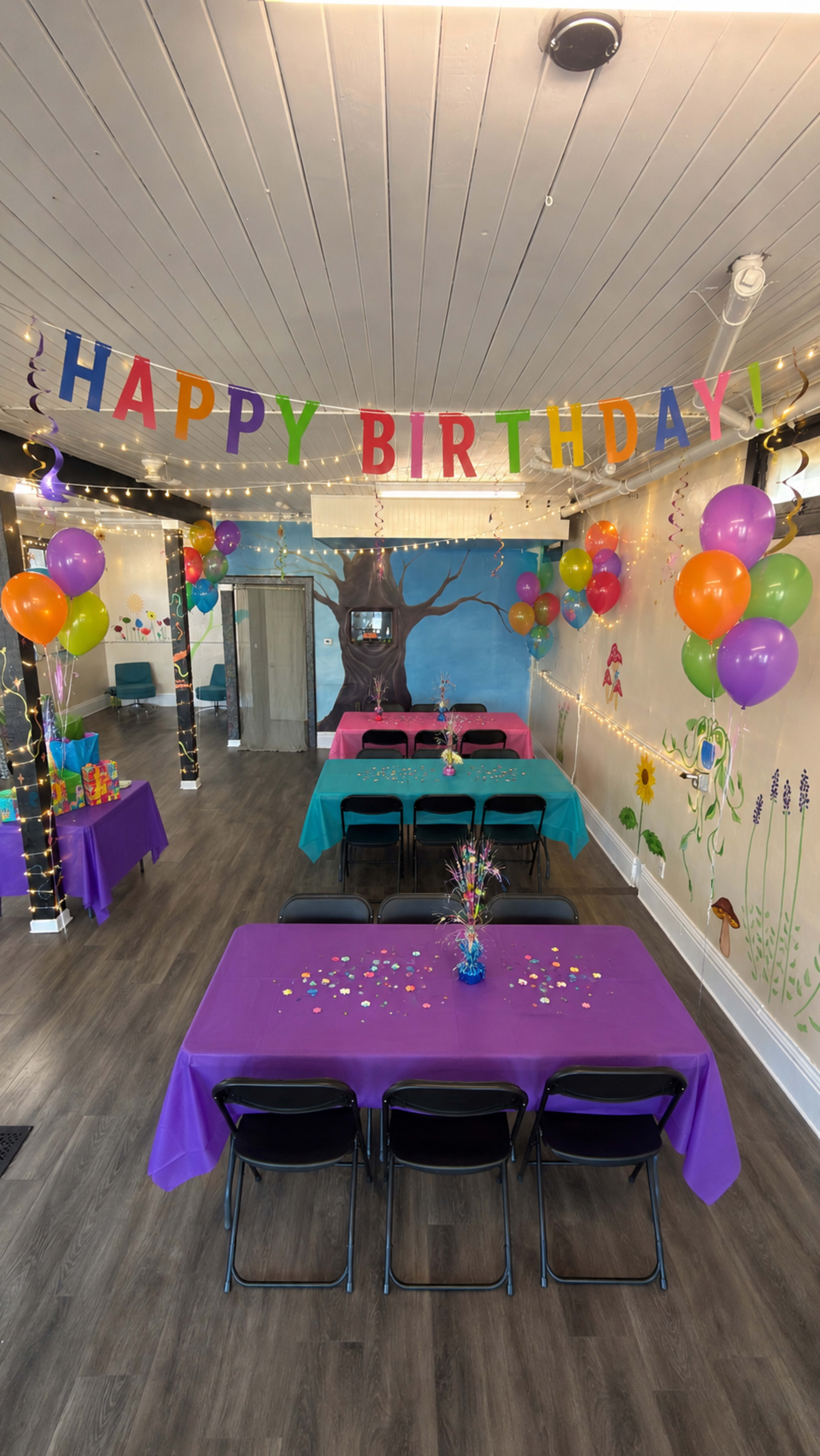 A brightly decorated birthday party venue features colorful tables, balloons, and a "HAPPY BIRTHDAY!" banner.