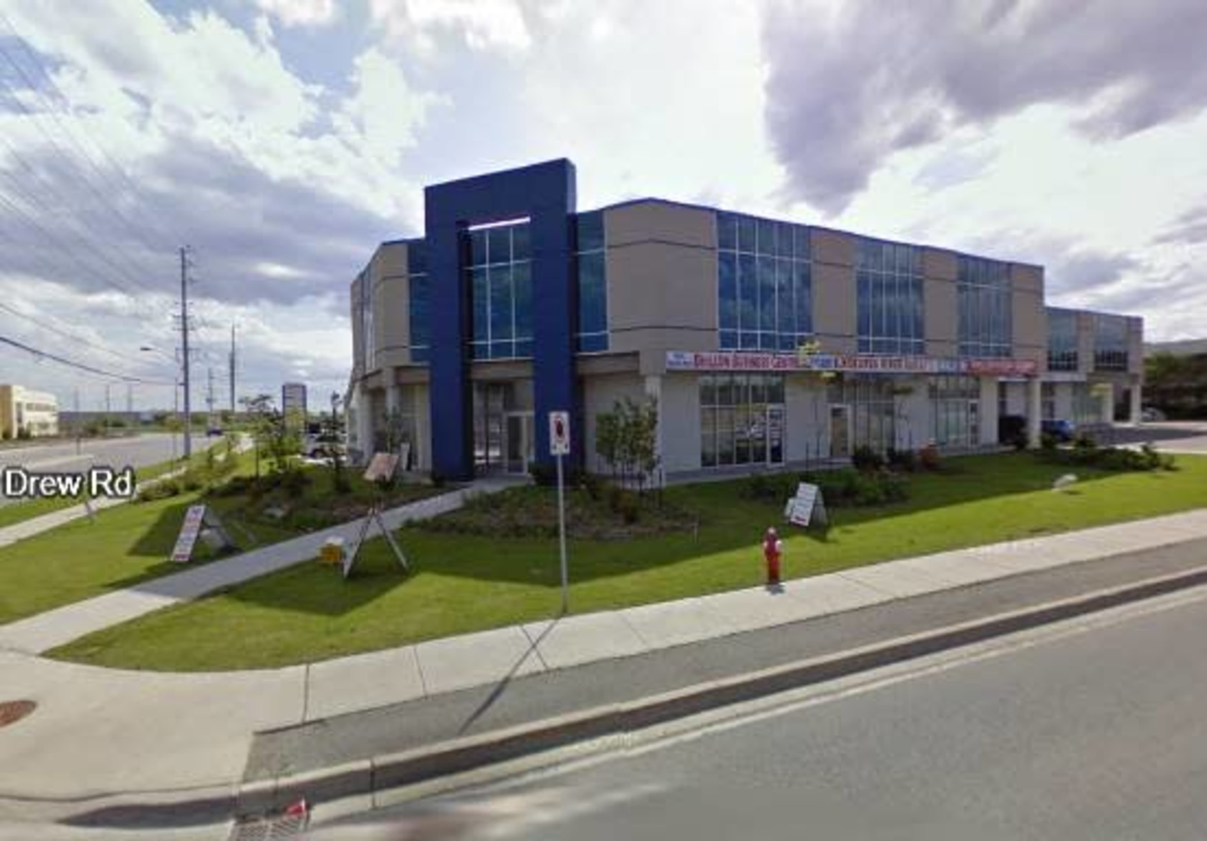 The image shows a modern commercial building with large windows and a landscaped grassy area, located at the corner of a street.