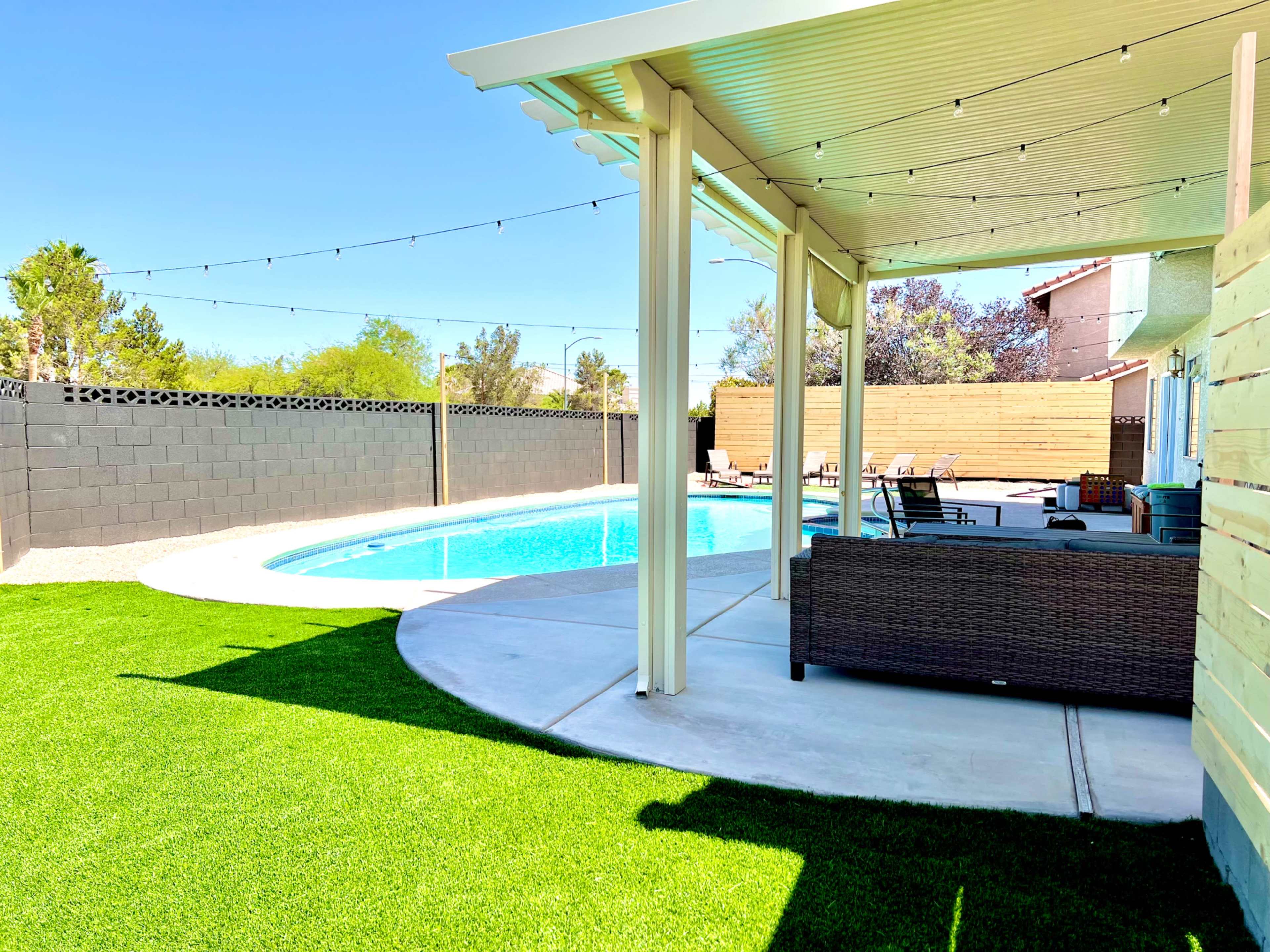 A backyard pool area featuring a swimming pool, a covered patio with furniture, and landscaped grass.