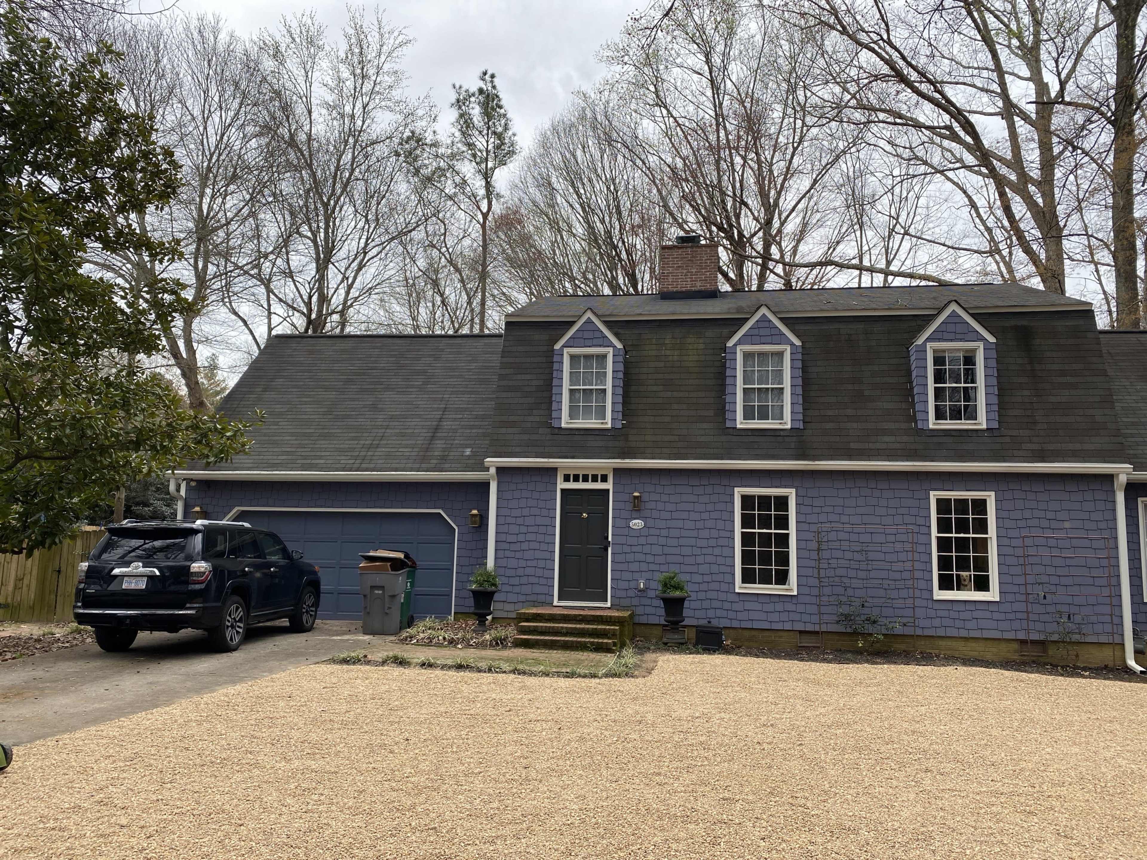 A two-story, blue house with a front porch and a gravel driveway is surrounded by trees.