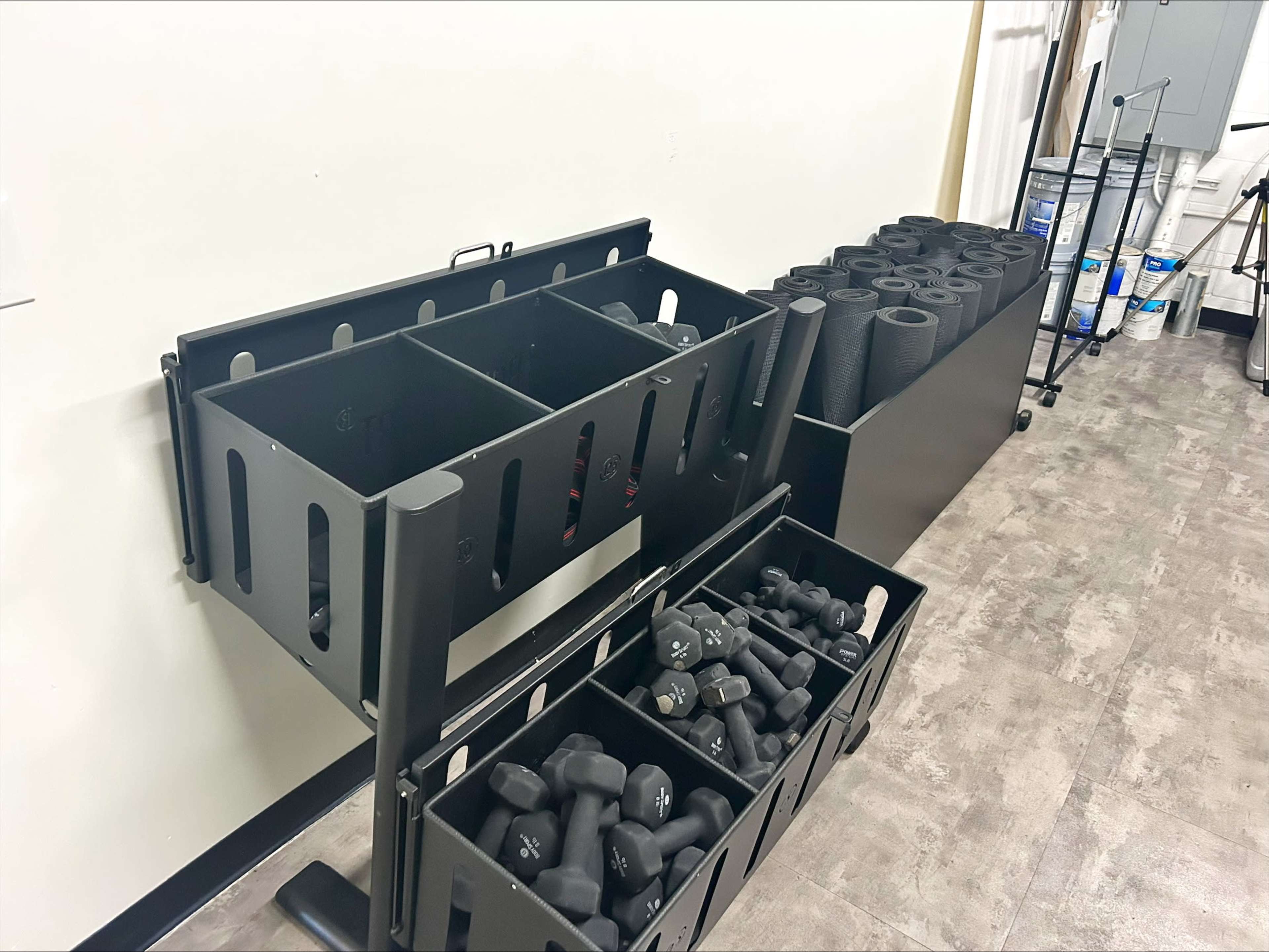 The image shows a black storage rack holding various dumbbells and foam rollers against a white wall in a gym or fitness area.