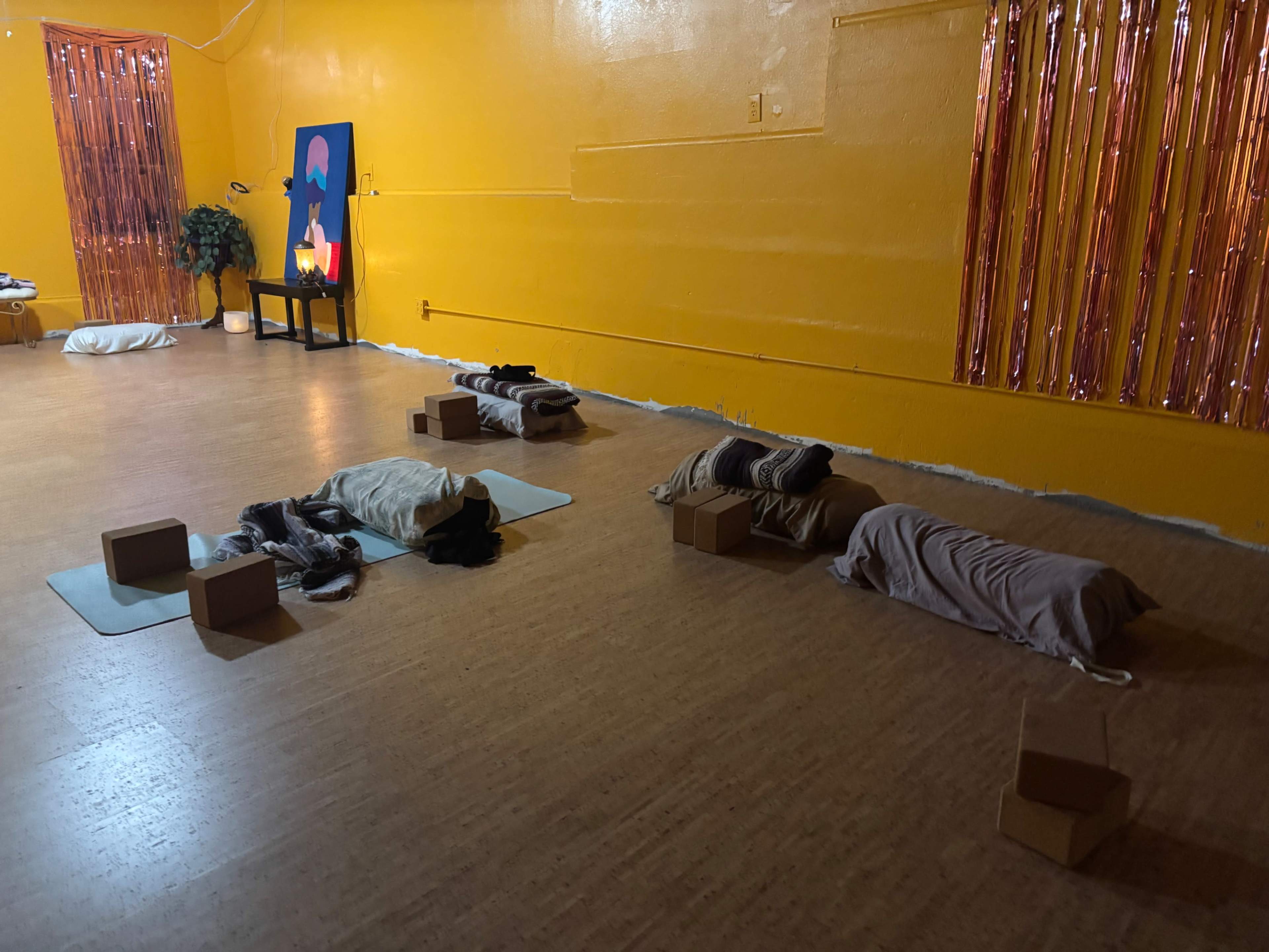 The image shows a brightly colored orange room with yoga mats and blankets arranged on the floor, alongside wooden blocks and a small table with a candle.