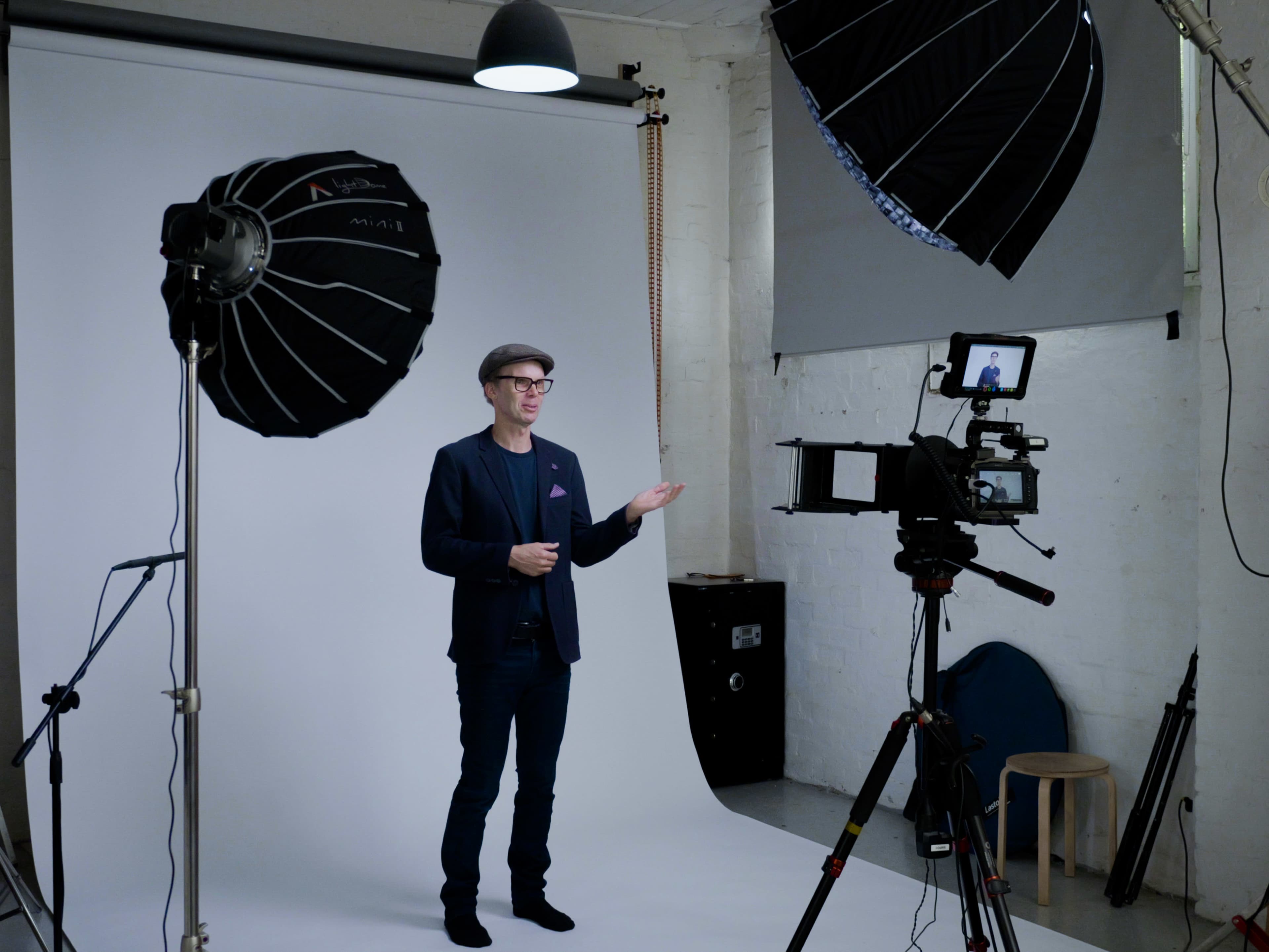 A man in a dark suit and hat stands in front of a white backdrop, gesturing towards a camera setup with large softbox lights on either side.