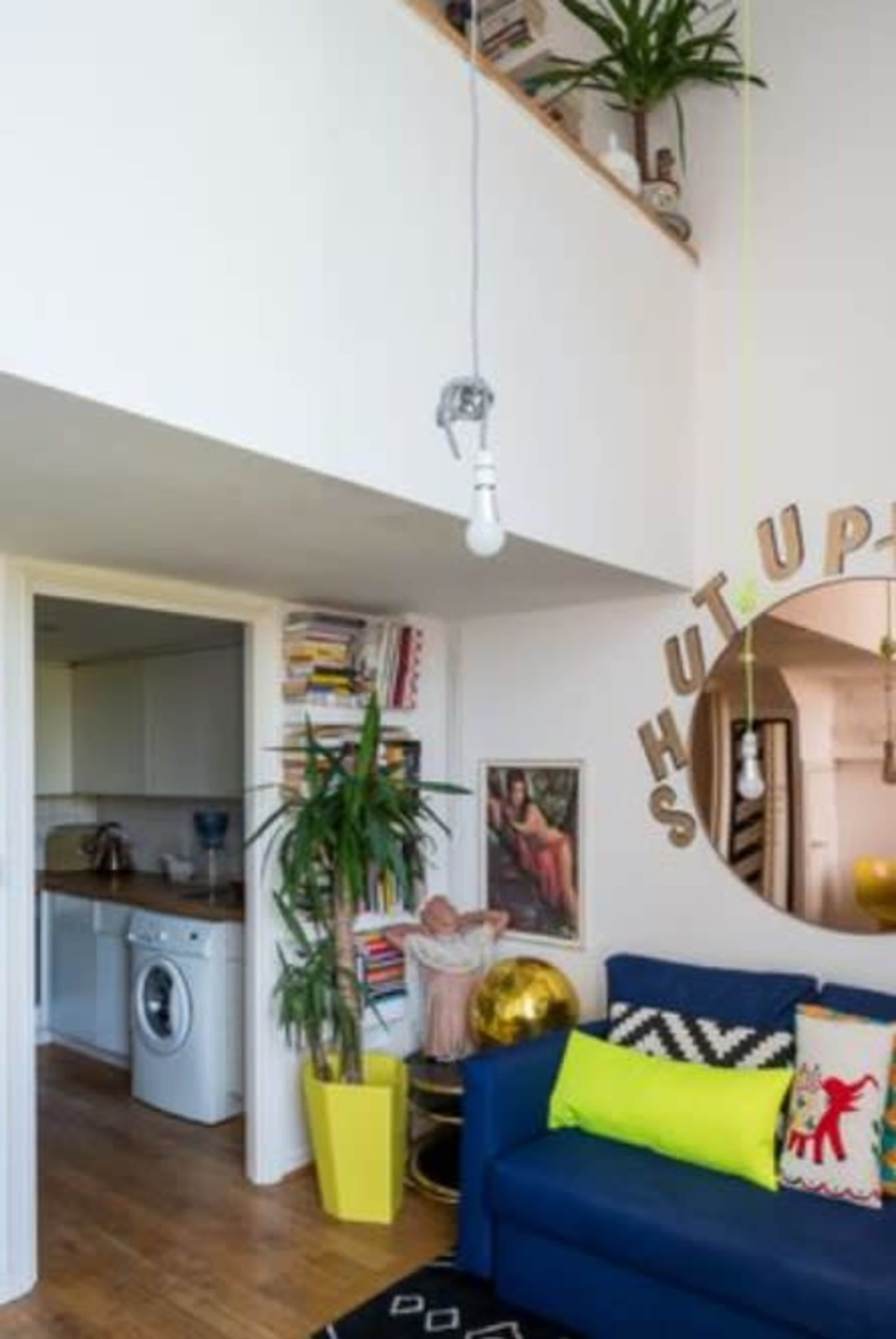 The image shows a modern living room with a blue sofa, a yellow plant pot, and a circular mirror on the wall, leading to a kitchen area in the background.