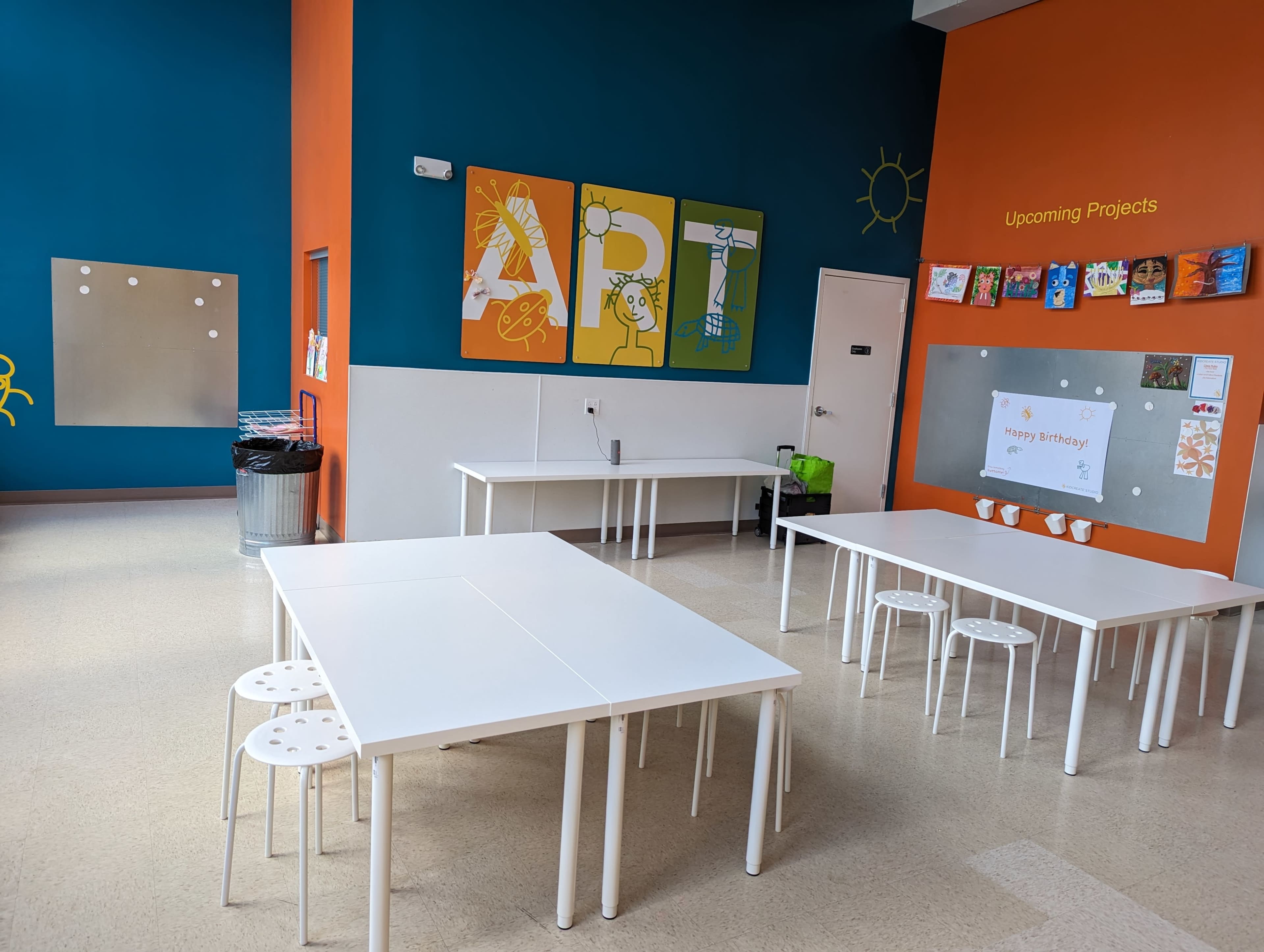 A bright, spacious art classroom with white tables and stools, colorful wall art, and a bulletin board displaying project information.