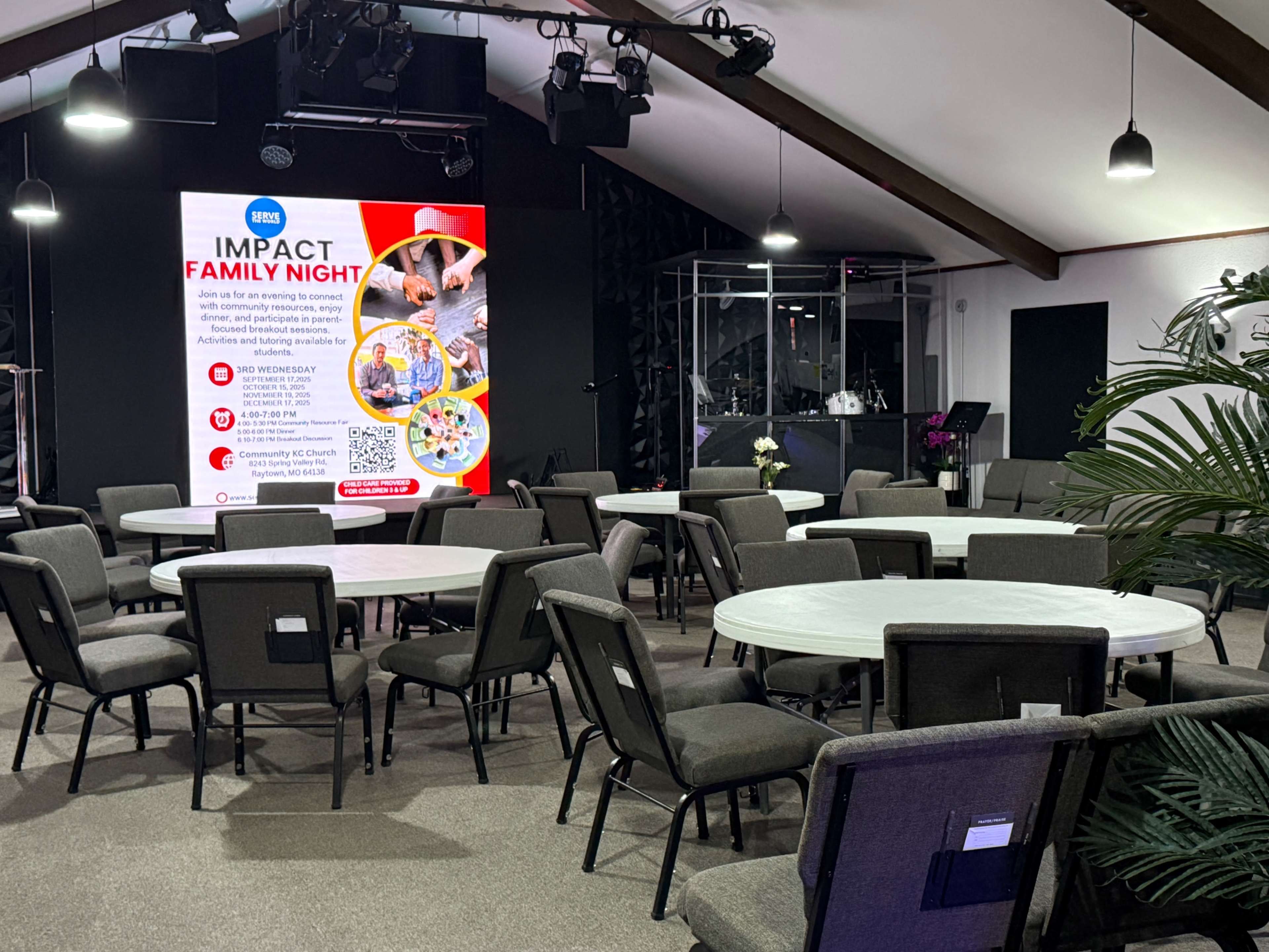 The image shows a spacious indoor venue with round tables and chairs arranged in a semi-circle, a stage area in the background, and a large screen displaying information about an event titled "Impact Family Night."