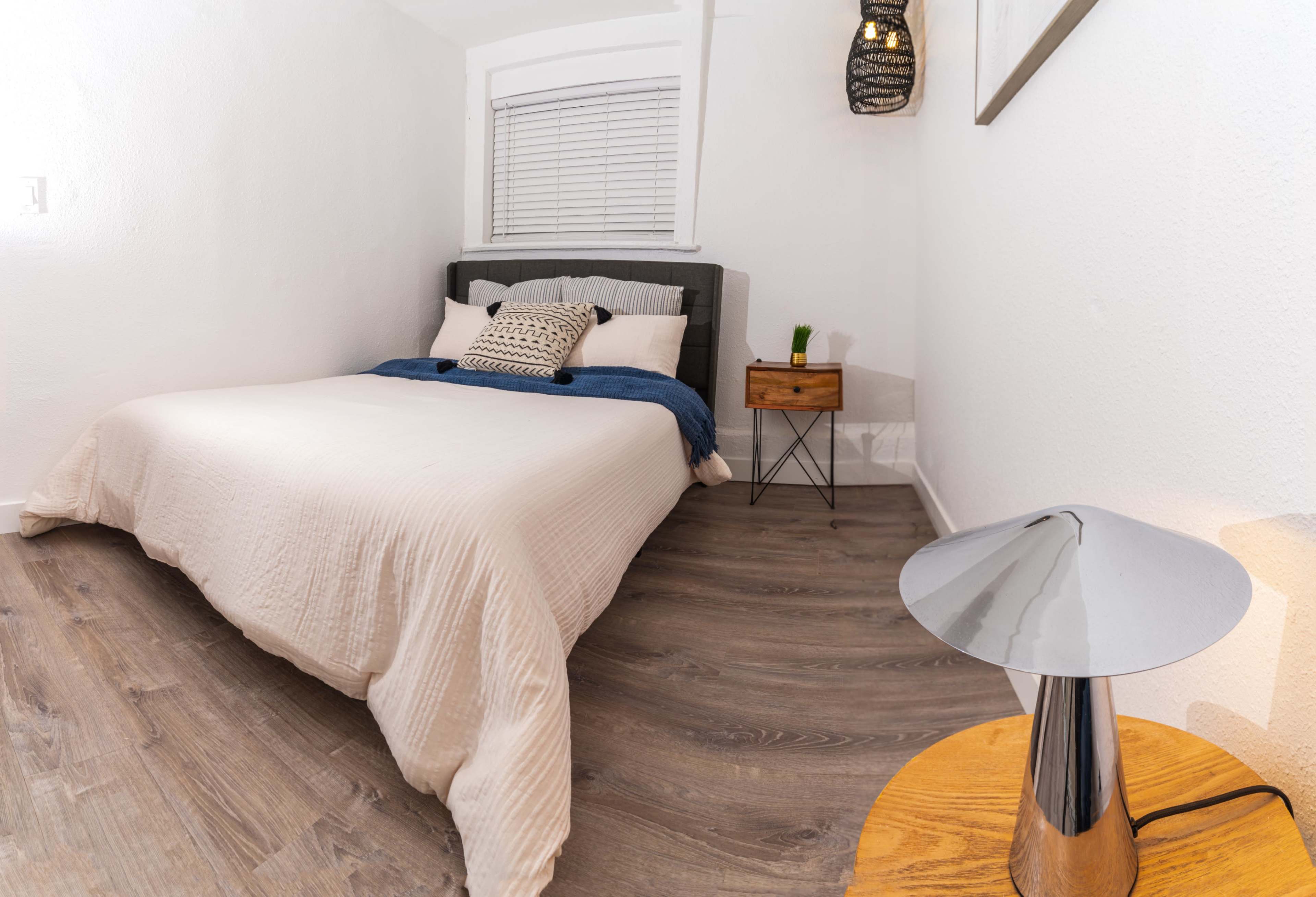 The image shows a neatly arranged bedroom featuring a bed with light bedding, a wooden nightstand with a green plant, and a modern lamp on the side.
