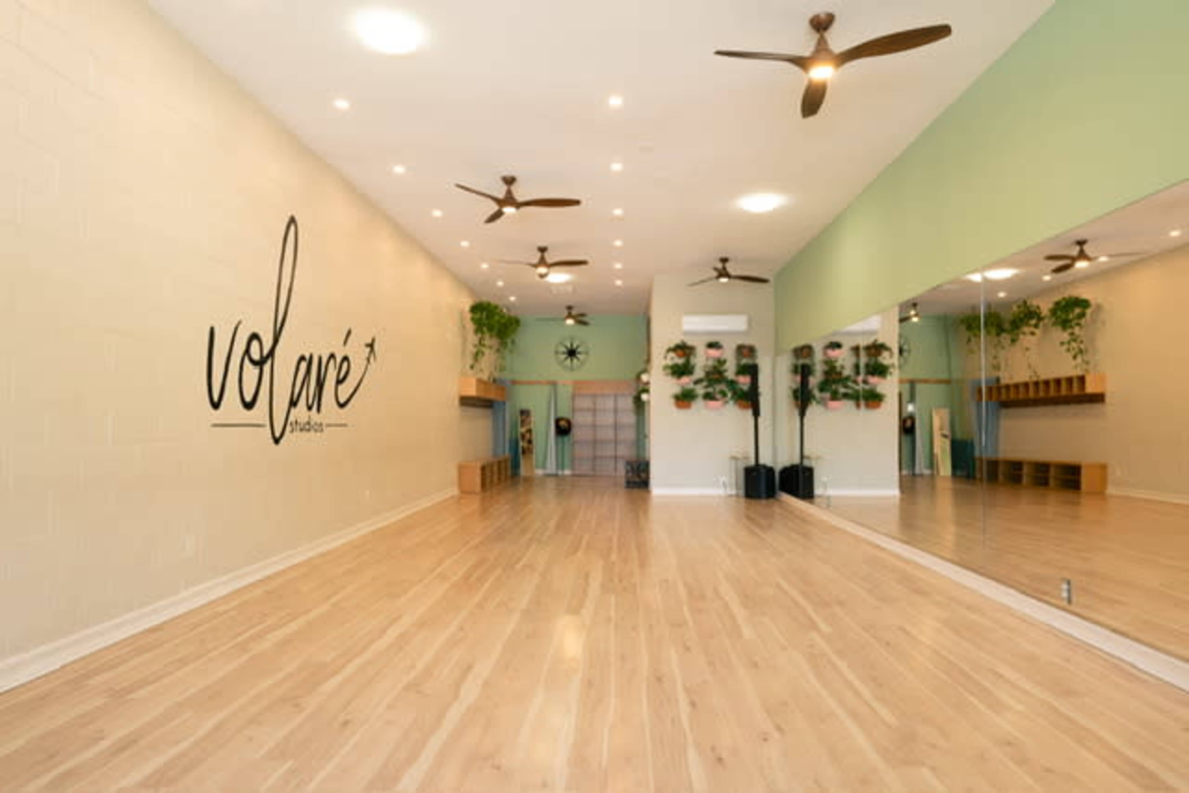 The image shows a spacious, well-lit yoga studio with wooden floors, green walls, and large mirrors, featuring potted plants and ceiling fans.