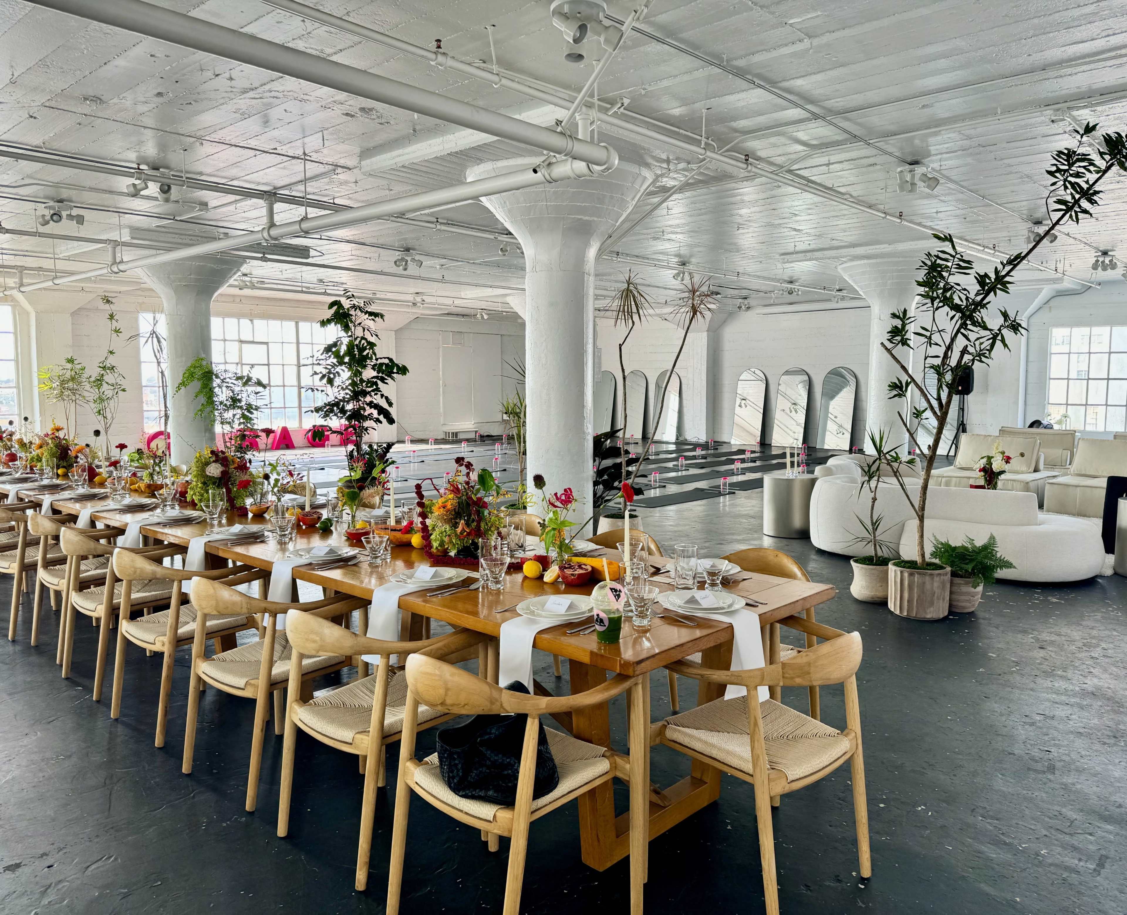 A long wooden dining table is set up in a spacious, brightly lit room with large windows, decorated with floral centerpieces and surrounded by modern seating.