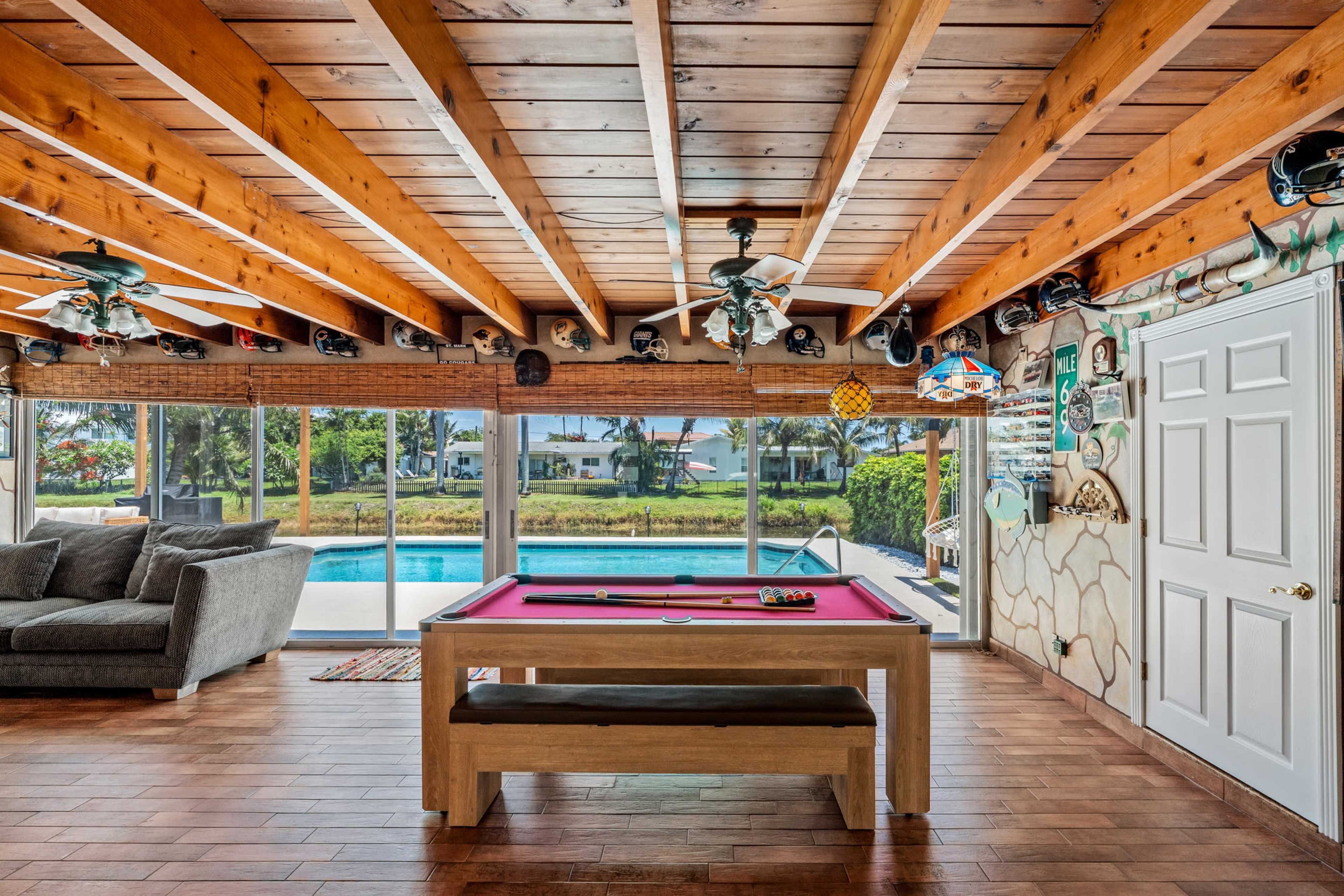 The image shows a spacious room with a pool table in the center, featuring wooden beams on the ceiling and a large window displaying a view of a pool and landscaped yard.