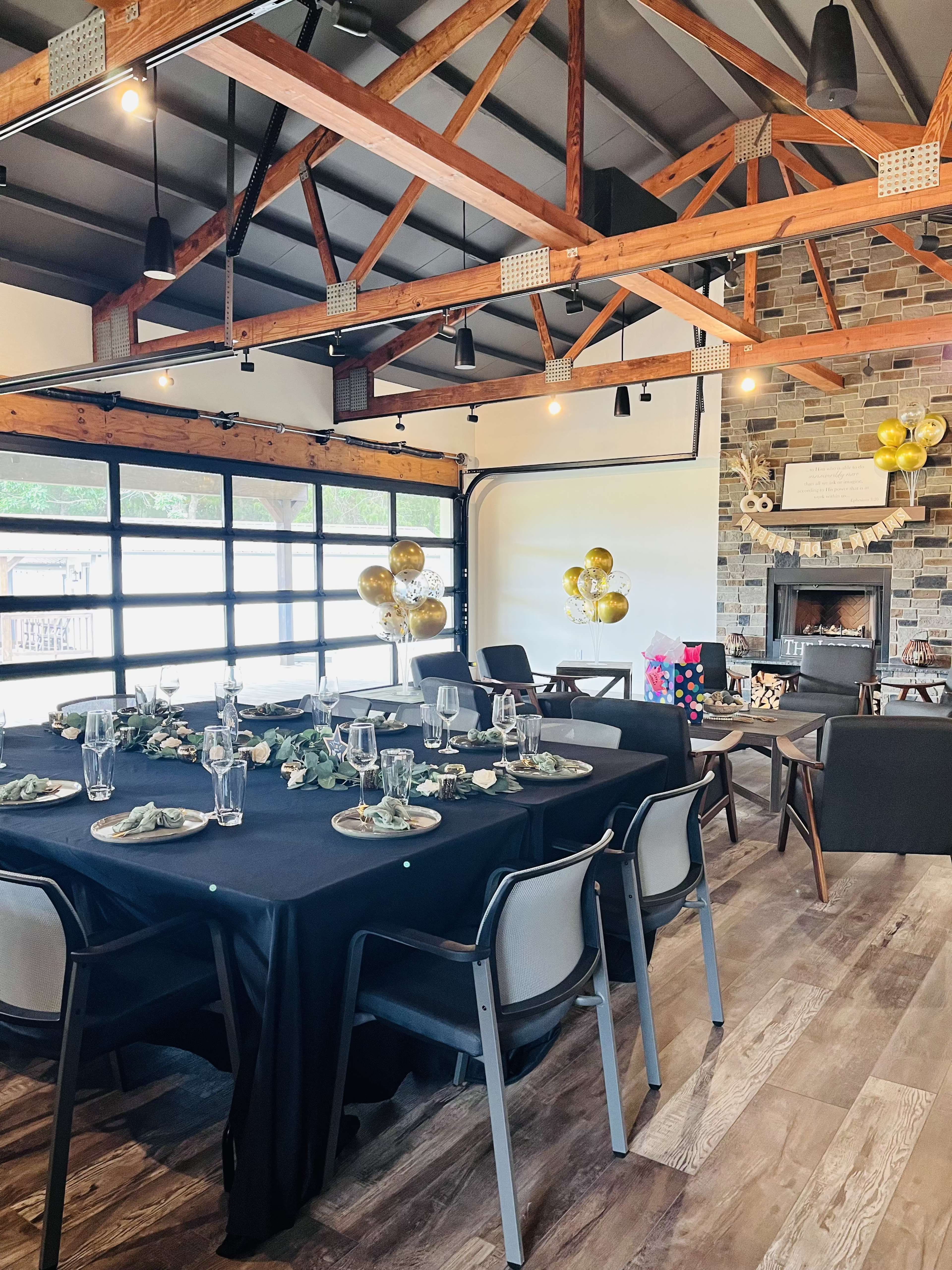 A decorated dining area with a black table set for a meal, surrounded by chairs and featuring a stone fireplace and balloons in the background.