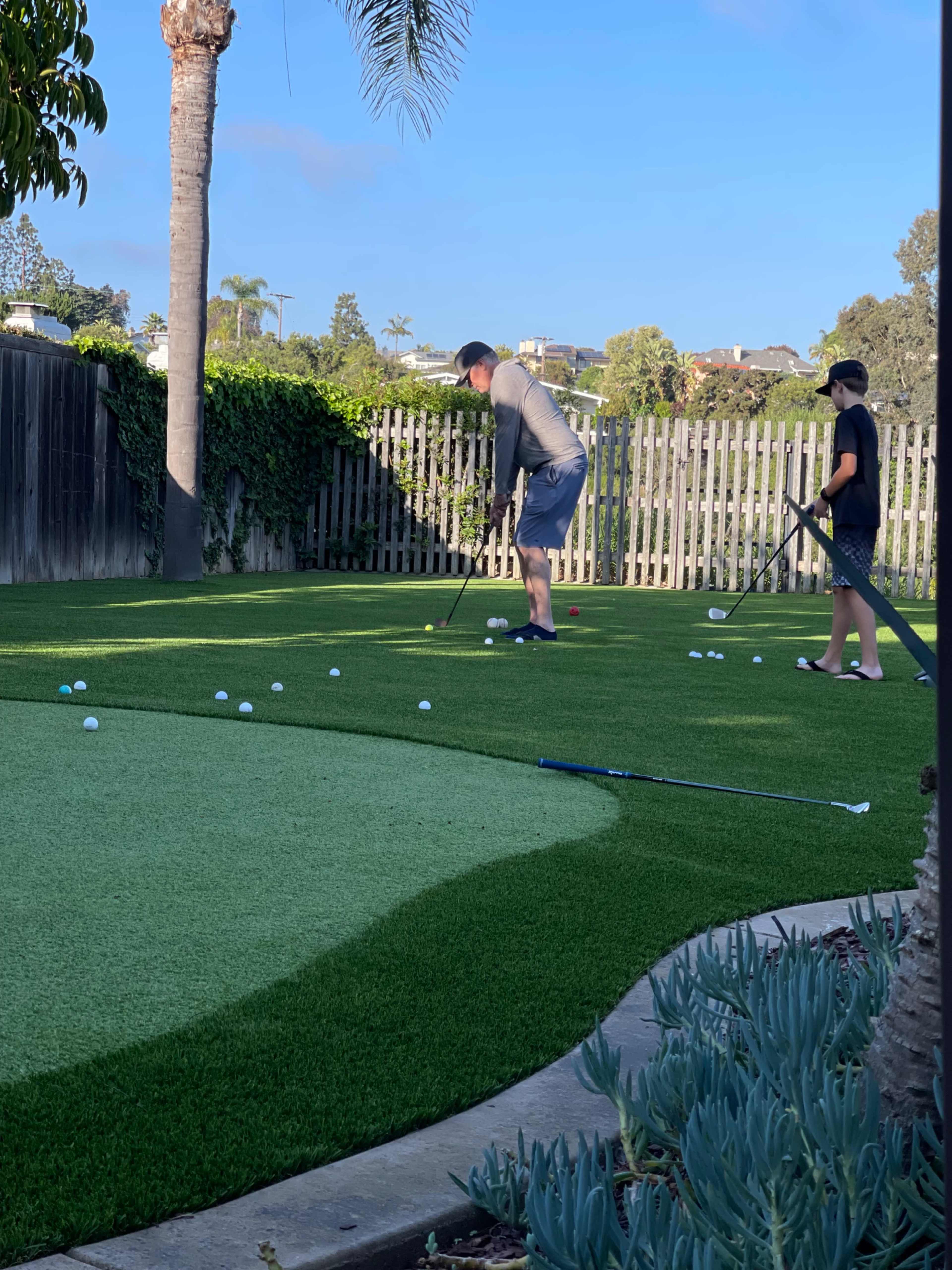 Two individuals practice golf putting on a synthetic grass green in a backyard setting, with a palm tree and houses visible in the background.