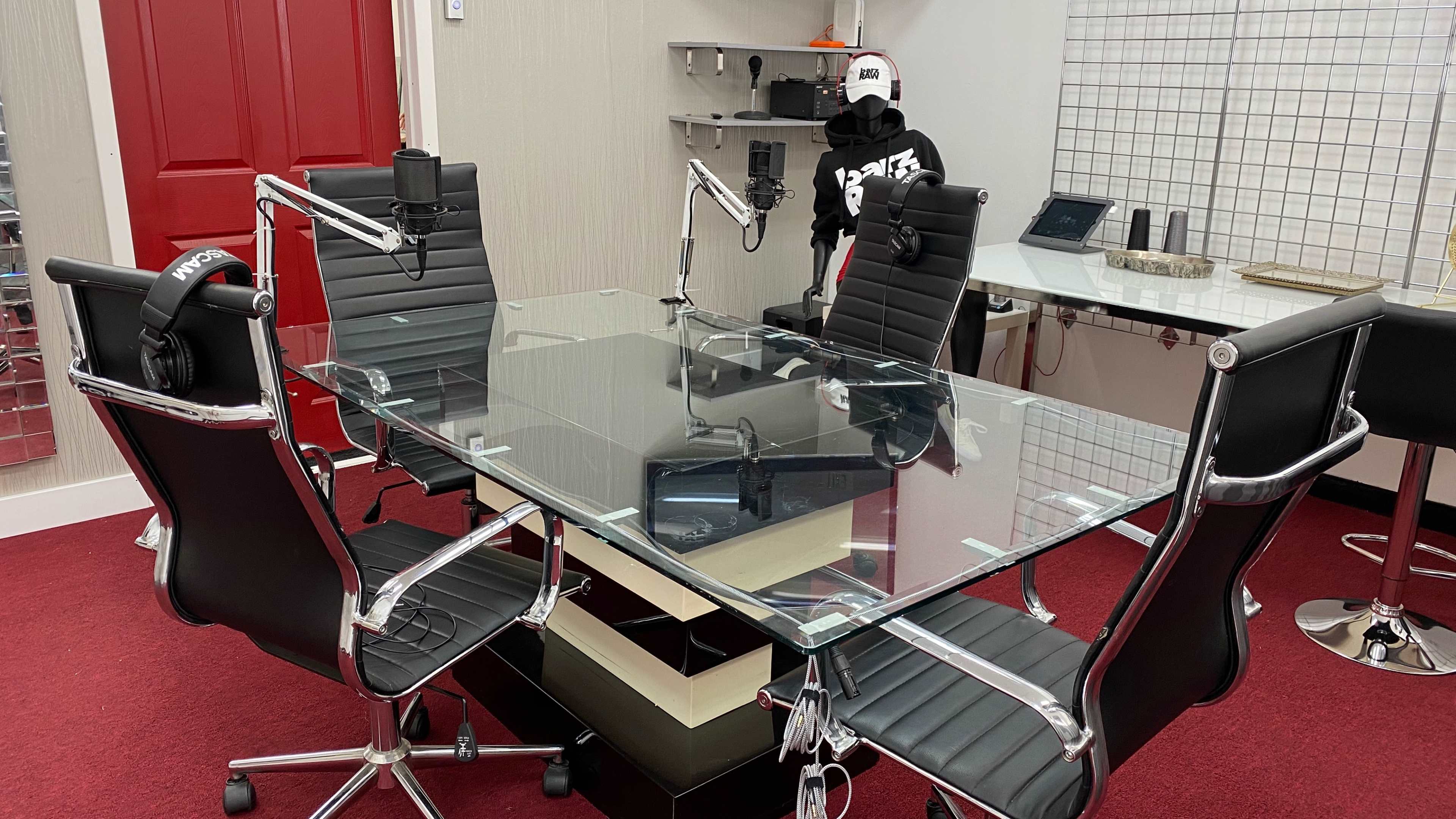A modern conference room features a glass table surrounded by black leather chairs, with microphones mounted on stands and a person in a black hoodie standing in the background.