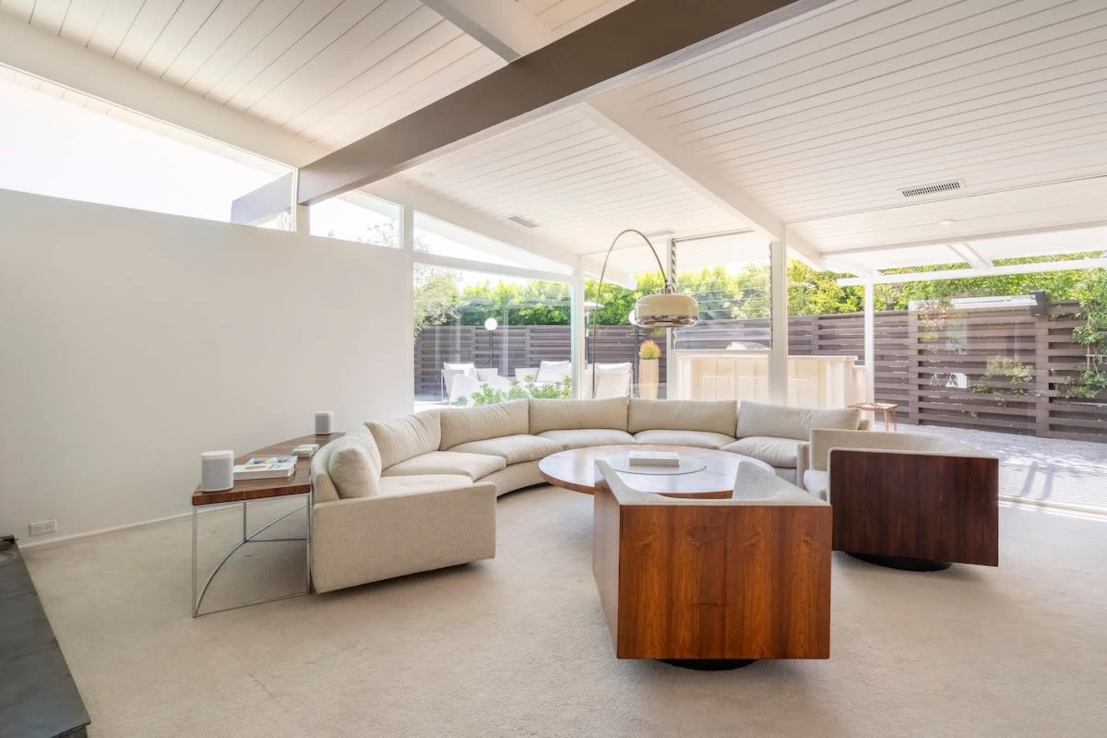 The image shows a modern living room with a curved beige sofa, two wooden side tables, a coffee table, and large windows overlooking a patio area.