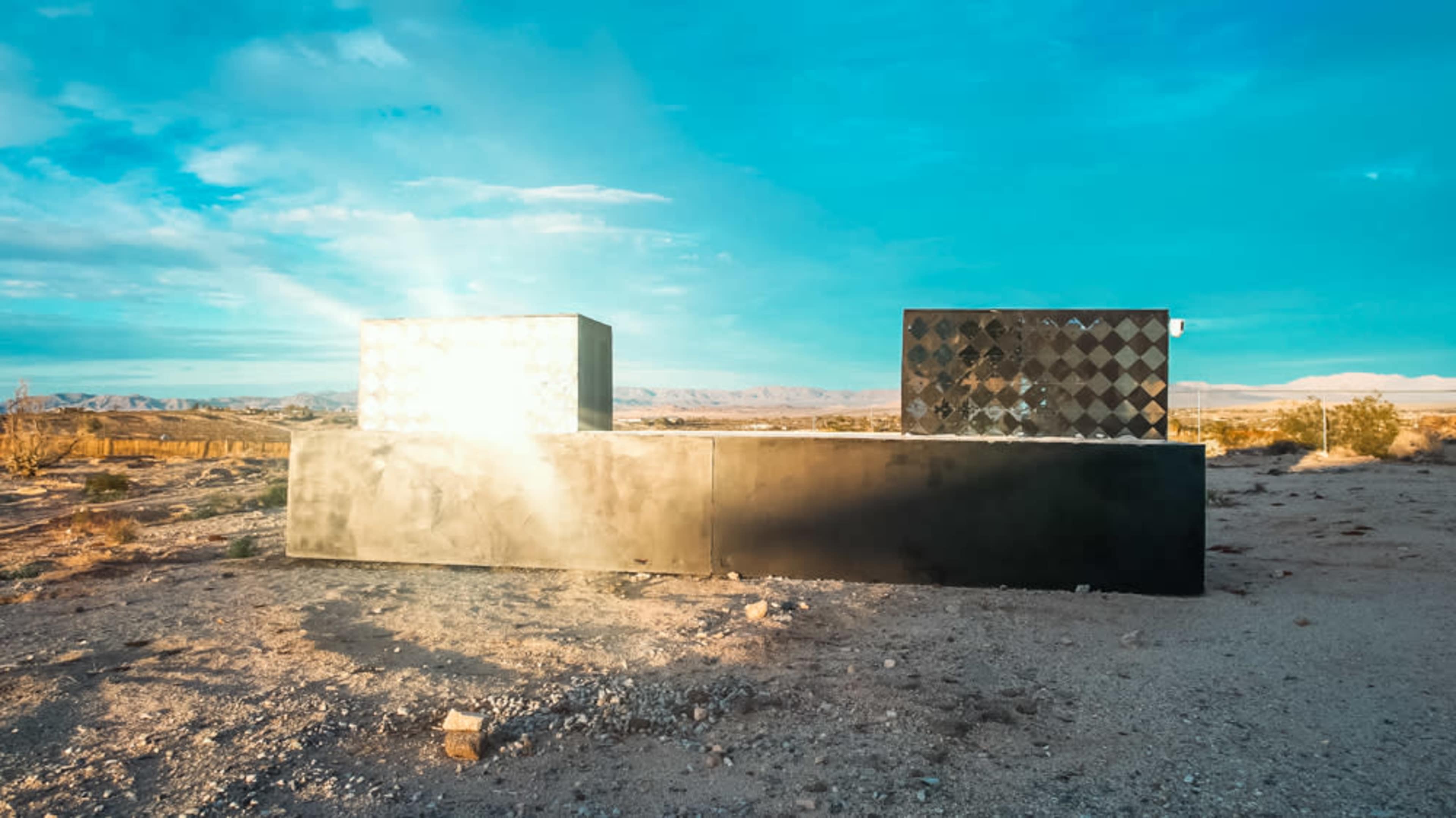 The image shows large geometric sculptures in a desert landscape under a blue sky.