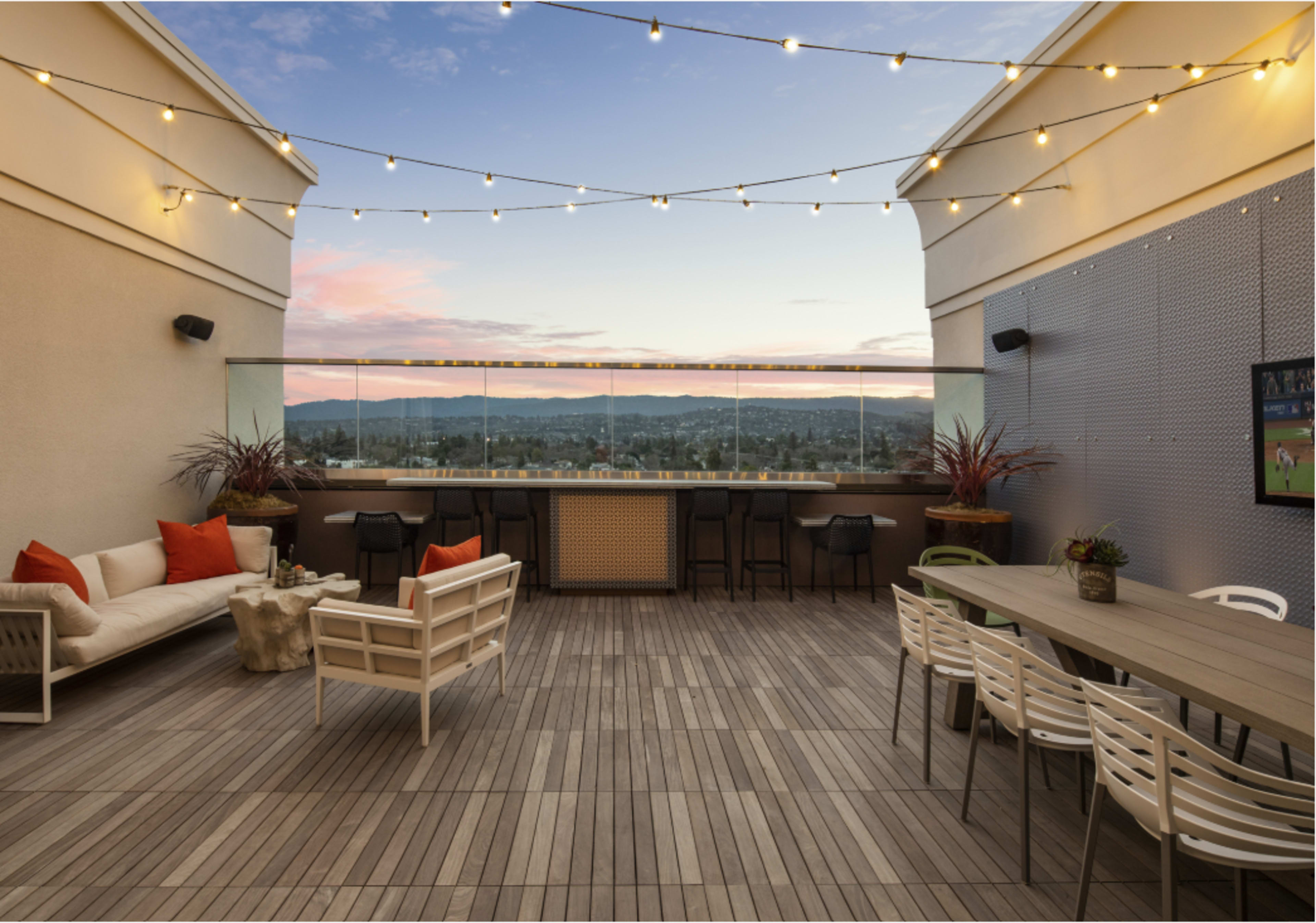 The image shows a rooftop patio with string lights, modern seating arrangements, and a view of distant mountains at sunset.