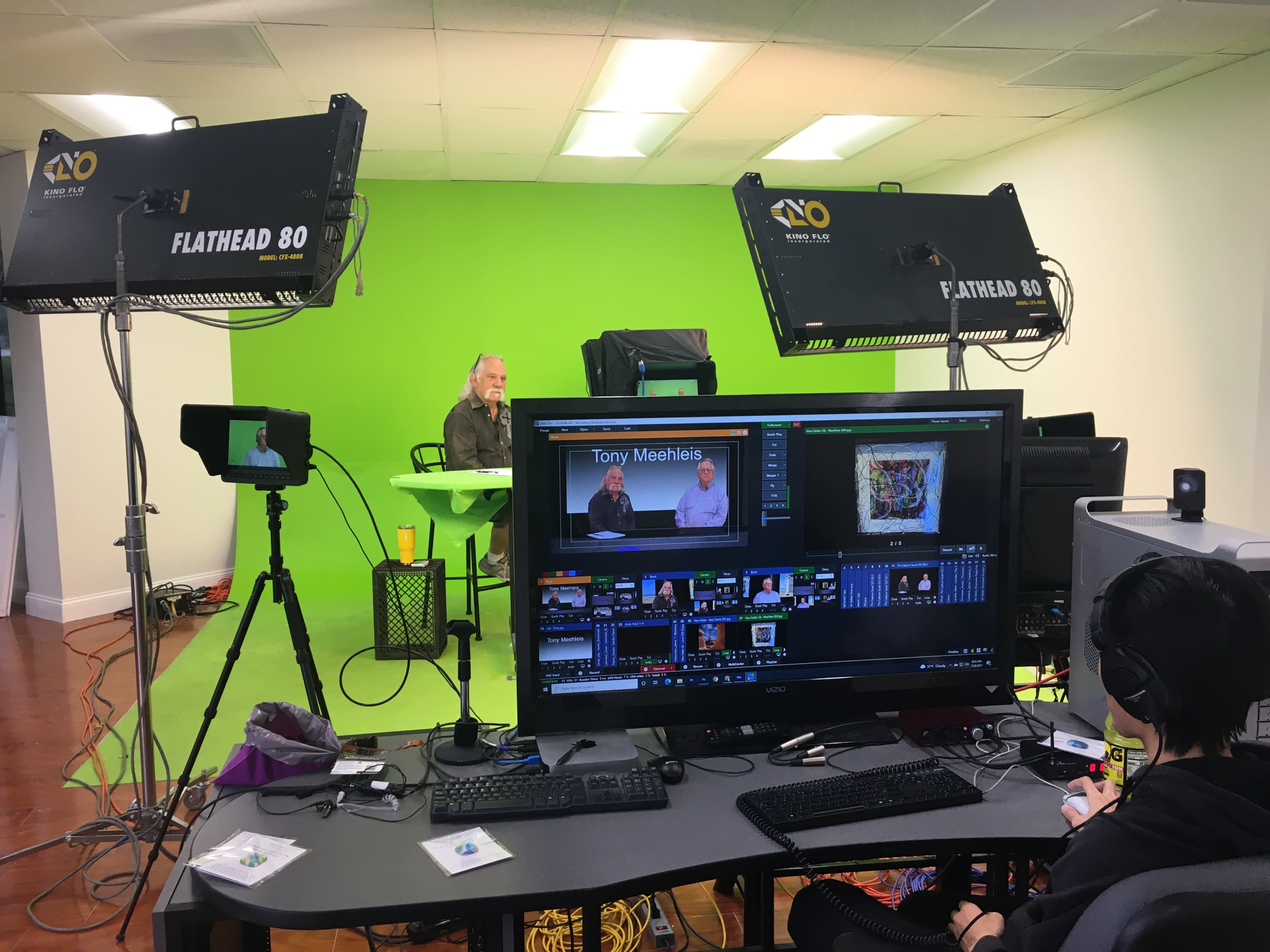 A video production setup is shown with two individuals seated at a table in front of a green screen, while a person operates a computer and equipment with multiple monitors.