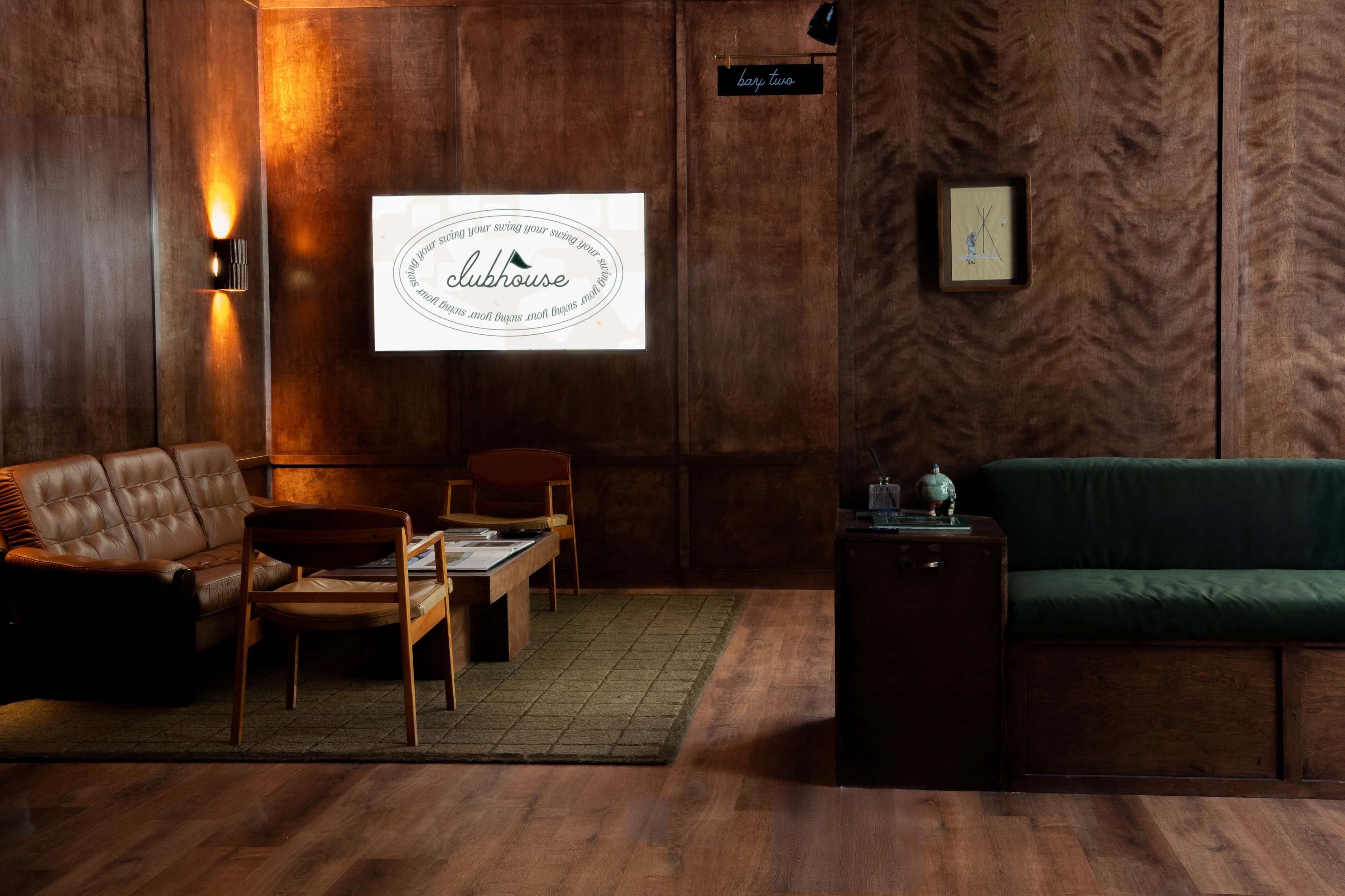 The image shows a cozy lounge area with wooden paneling, featuring brown leather sofas, a coffee table, and a green couch against a wall displaying a logo for "clubhouse."