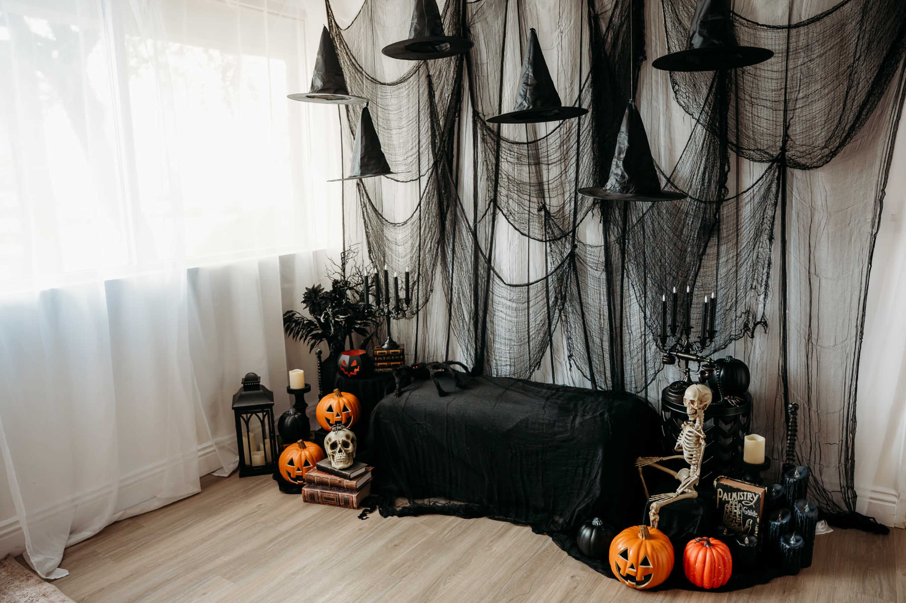 The scene depicts a corner of a room decorated for Halloween, featuring a black draped couch, hanging witch hats, cobwebs, pumpkins, a skeleton, and various candles.