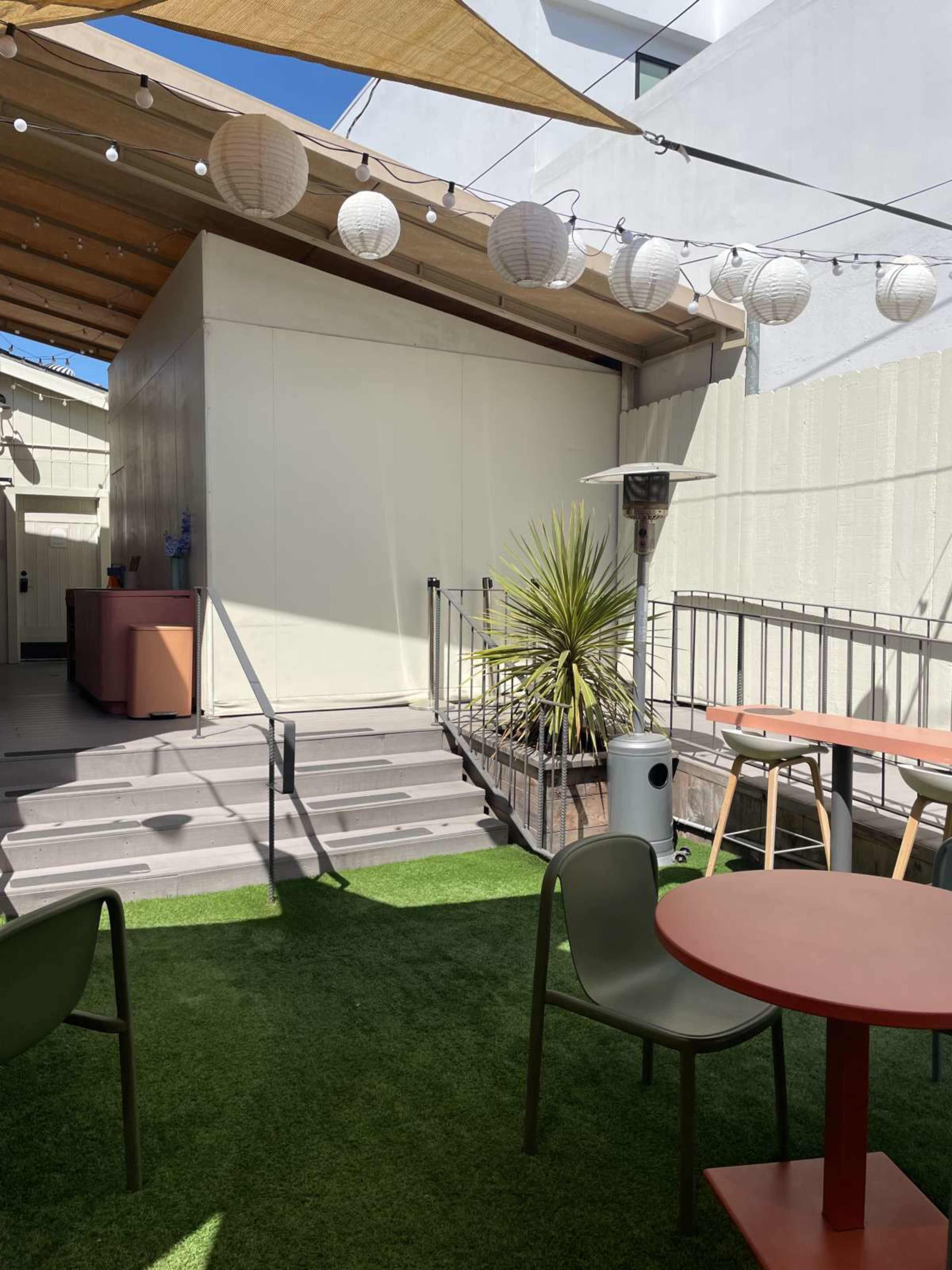 The image features a patio area with green artificial grass, a few tables, chairs, and decorative lanterns overhead, along with a potted plant and a heating fixture.