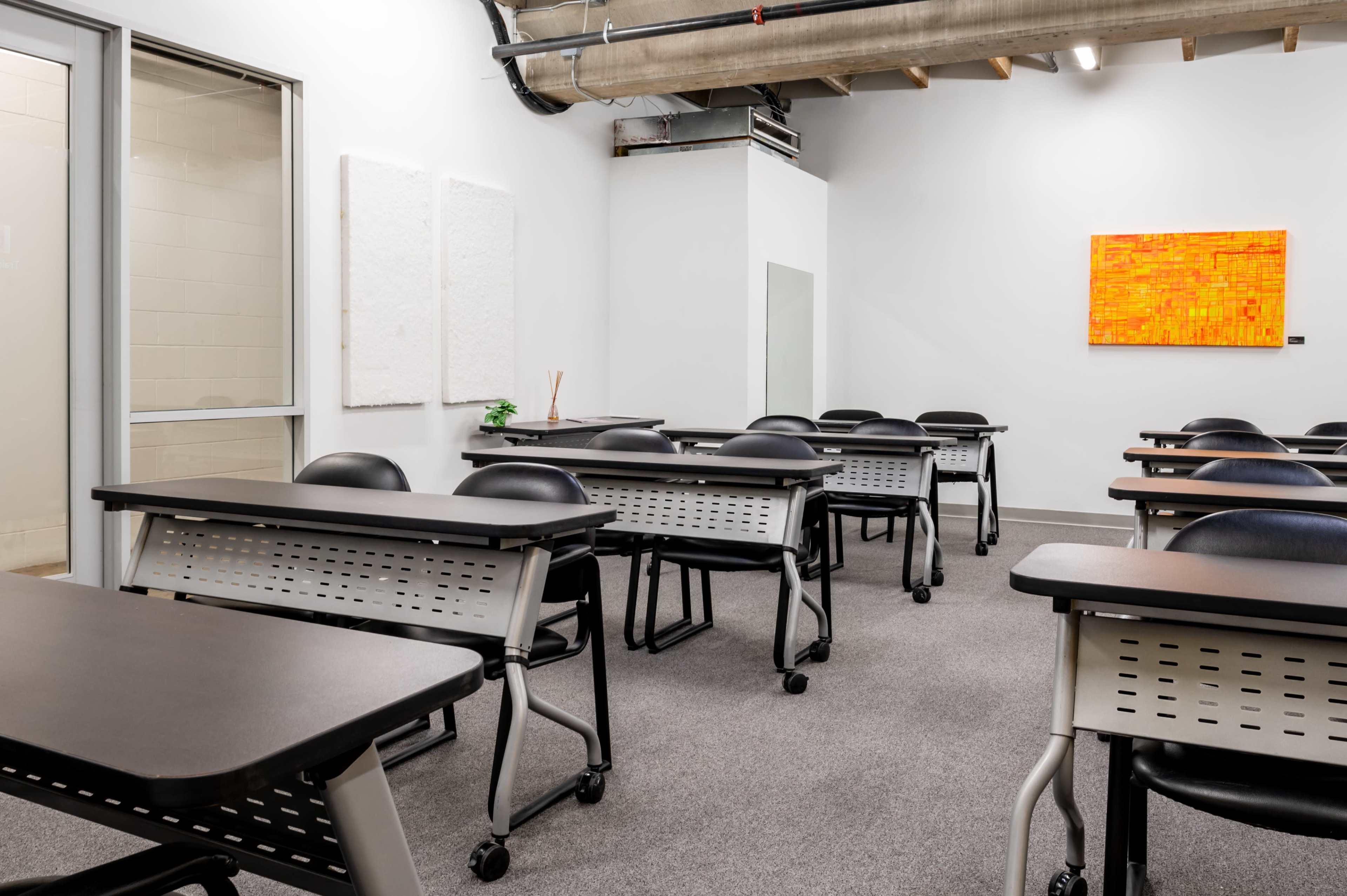 An empty classroom features rows of black chairs and desks arranged on a gray carpet, with a colorful artwork hung on the wall.